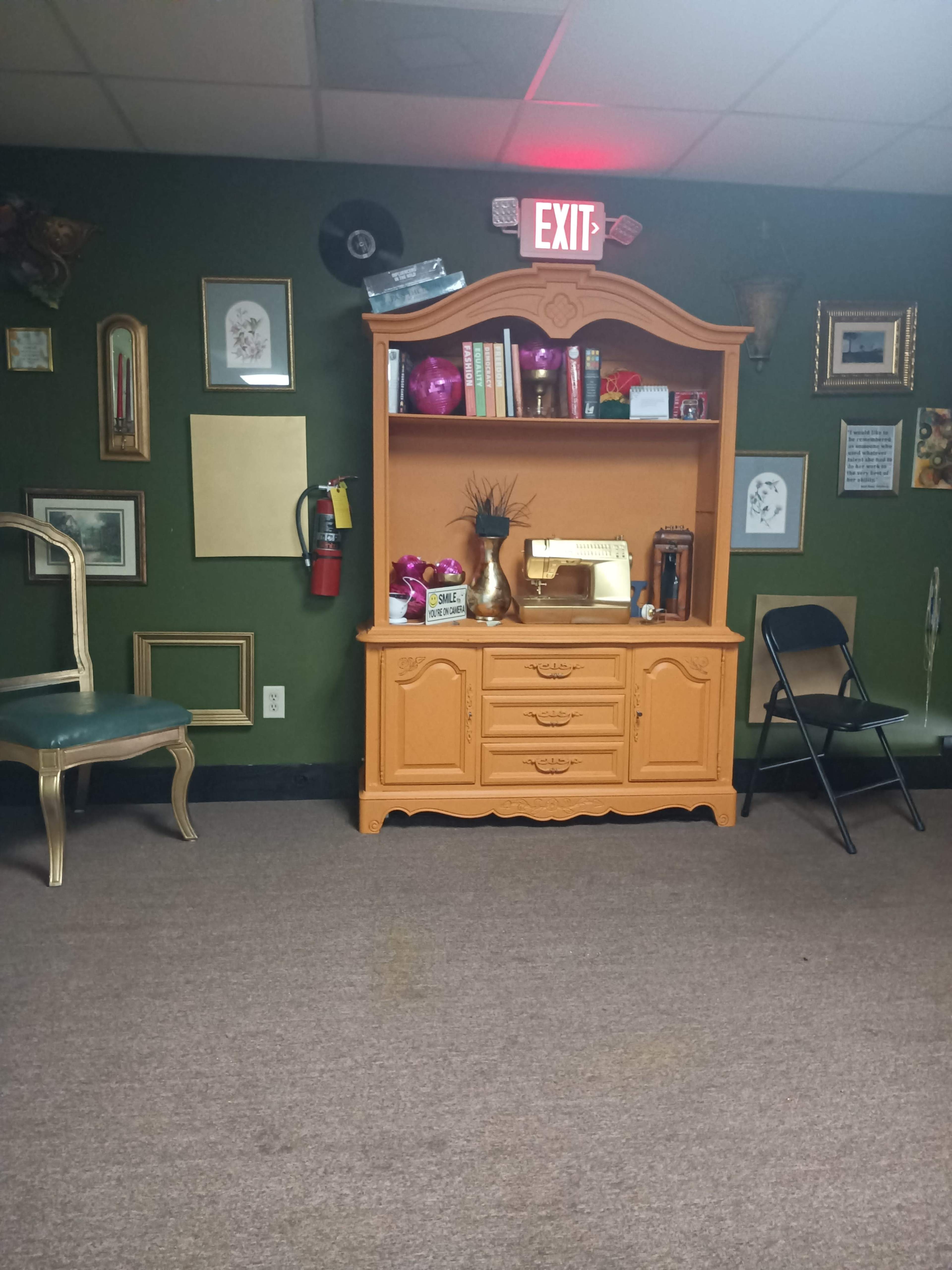 The image shows a brown dresser with decorative items on top, positioned against a green wall adorned with various framed artworks and an exit sign overhead.