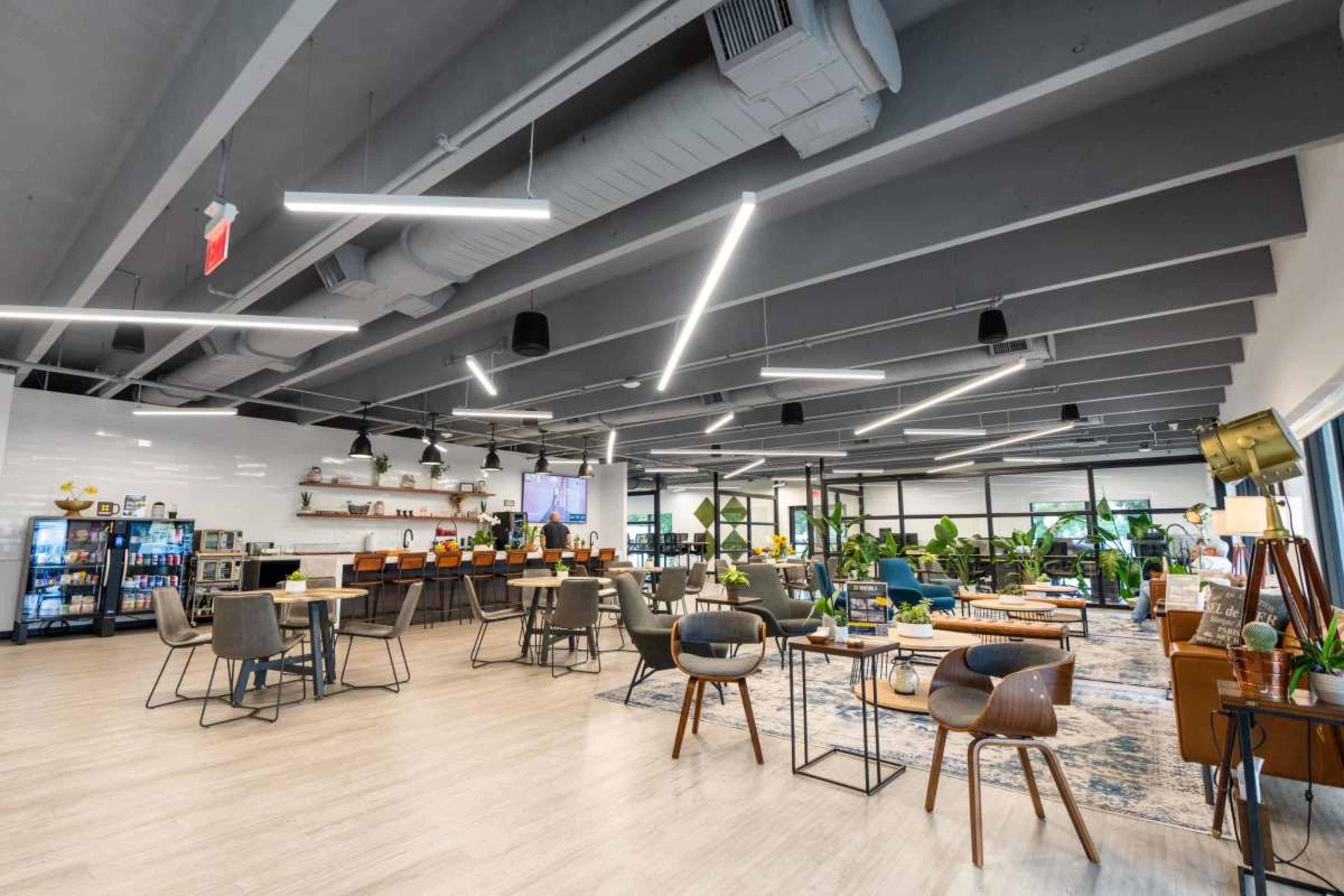 The image shows a modern open office space featuring a casual seating area, a kitchen with bar seating, and green plants throughout the room.