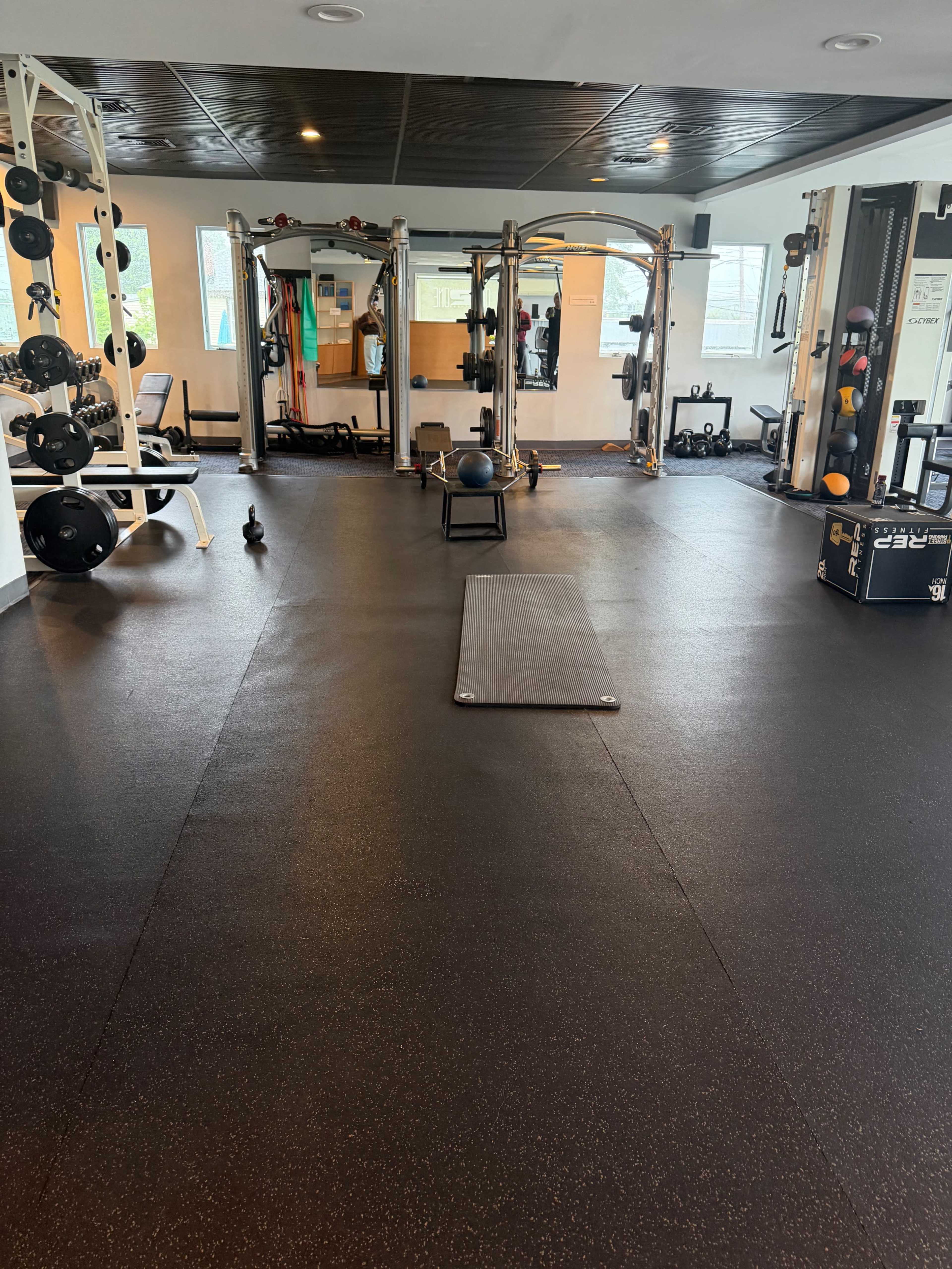 The image shows an empty exercise gym equipped with various weightlifting machines, free weights, a yoga mat, and mirrors along the walls.
