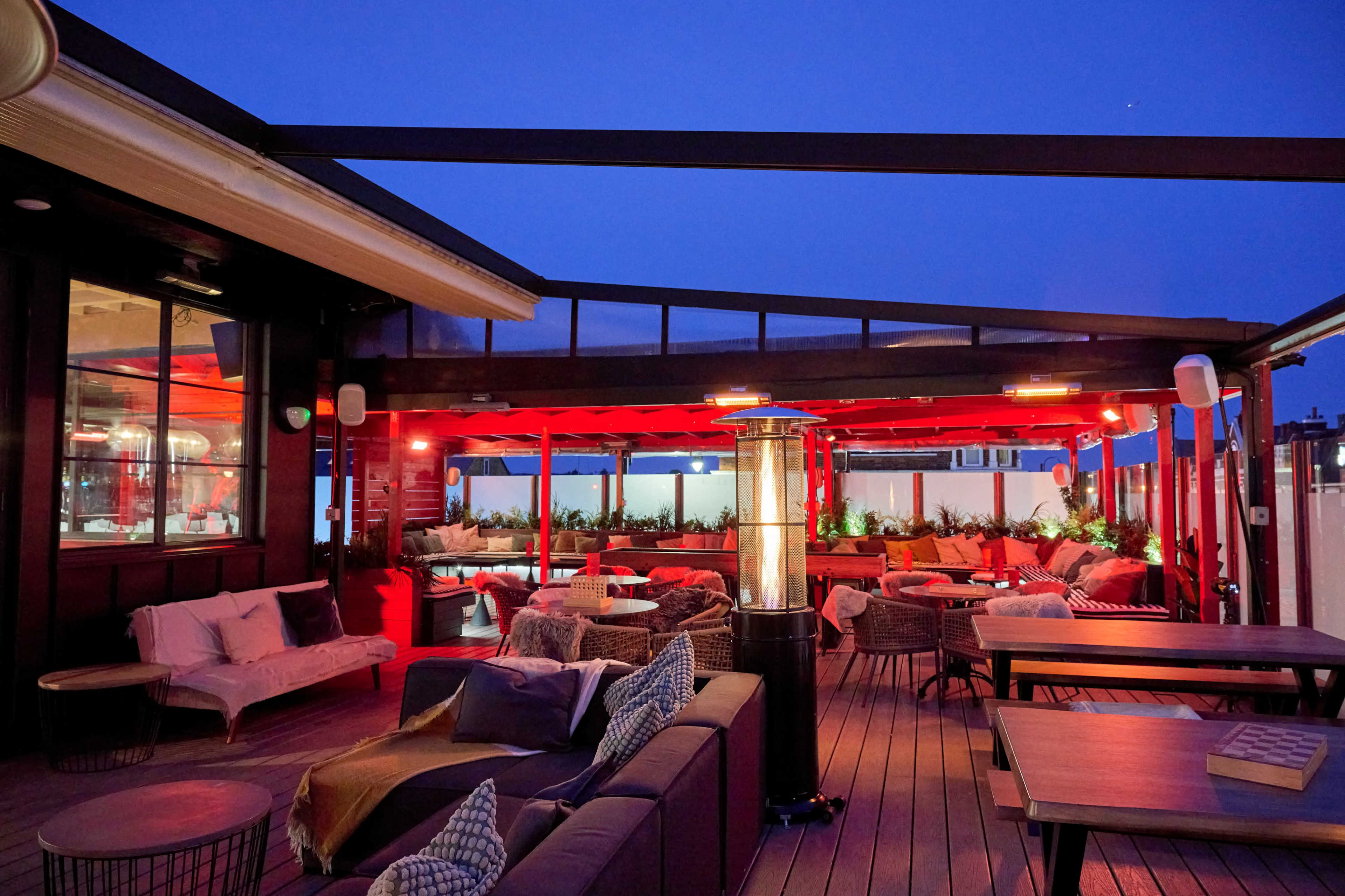 The image shows a rooftop terrace featuring modern seating arrangements, ambient red lighting, and plants, set against a twilight sky.