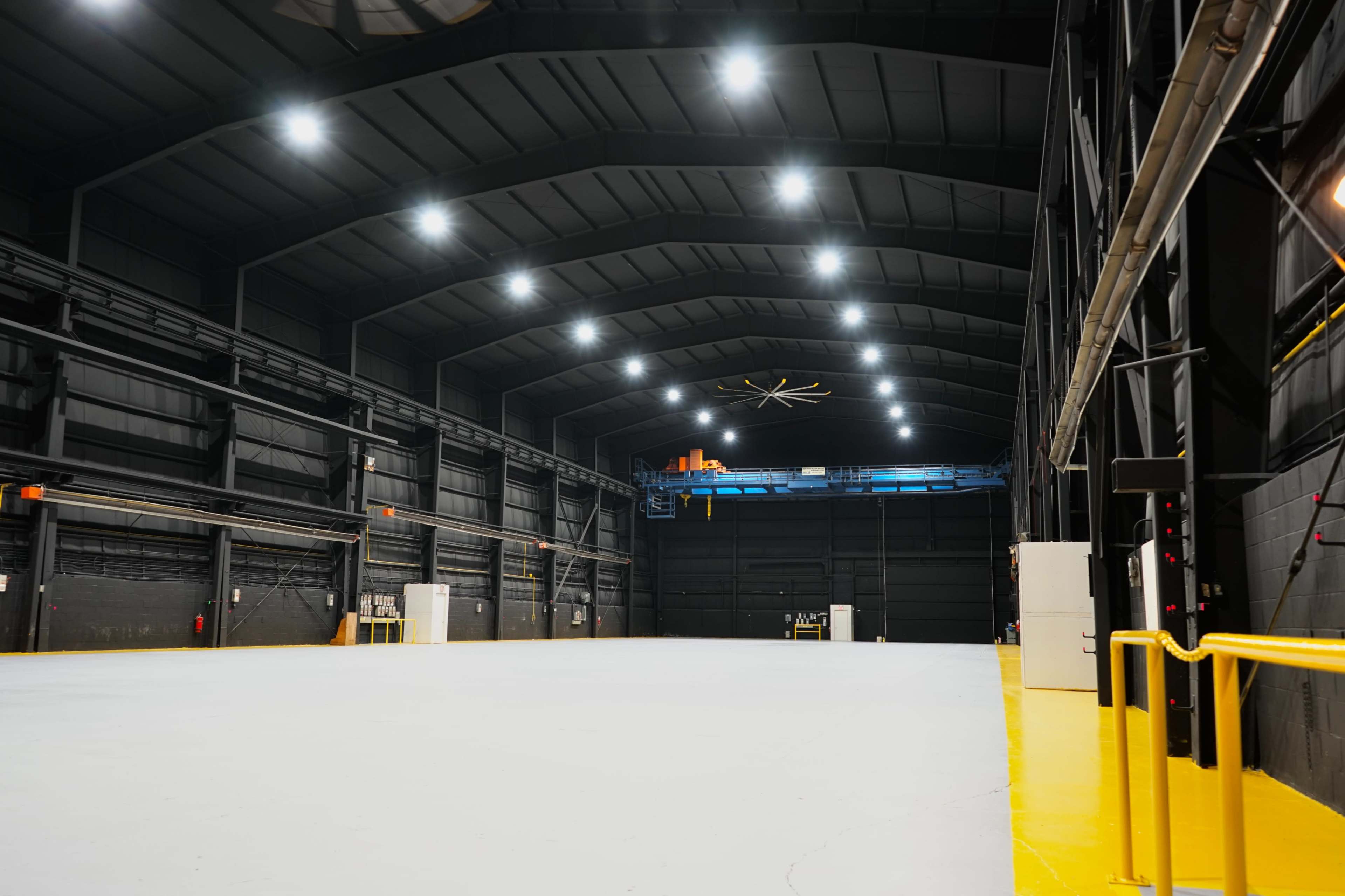 The image shows a large, empty industrial warehouse with a high ceiling, bright overhead lights, and a wide, open floor space.