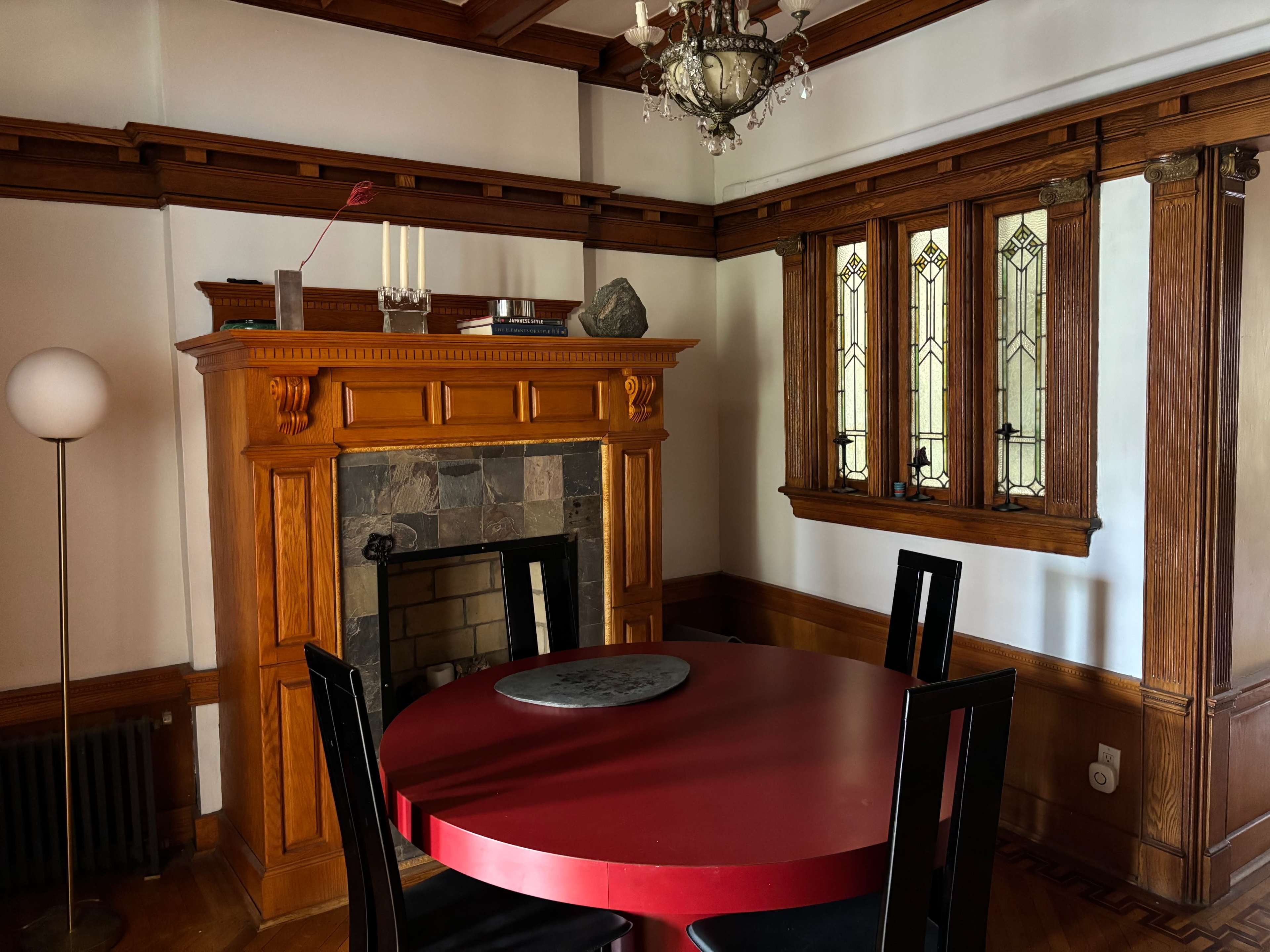 The image shows a cozy dining area with a red circular table, black chairs, an ornate wooden fireplace, and stained glass windows.