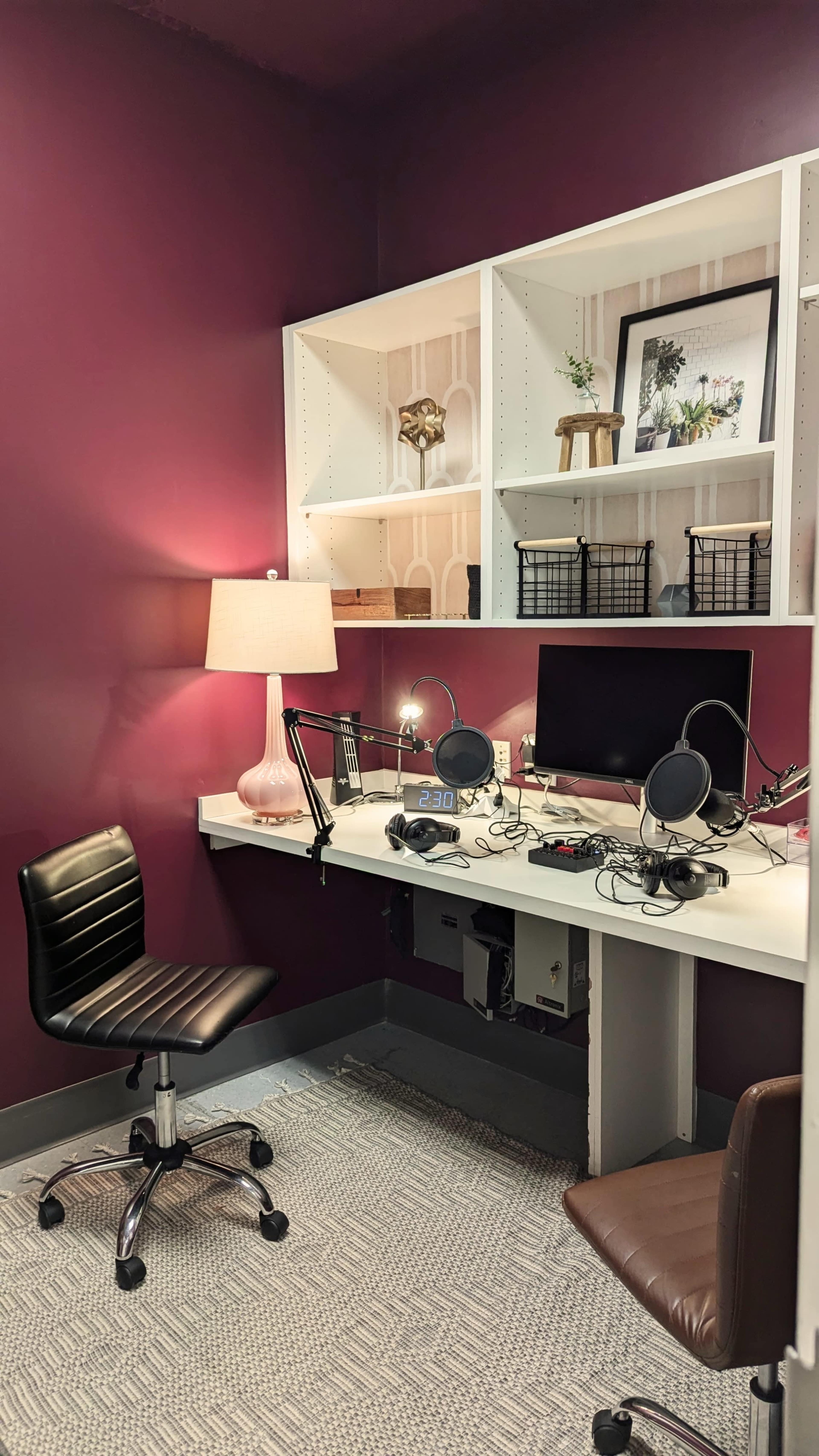 The image shows a small recording studio with a desk, two microphones, a computer monitor, and two chairs, set against a deep purple wall with white shelving.