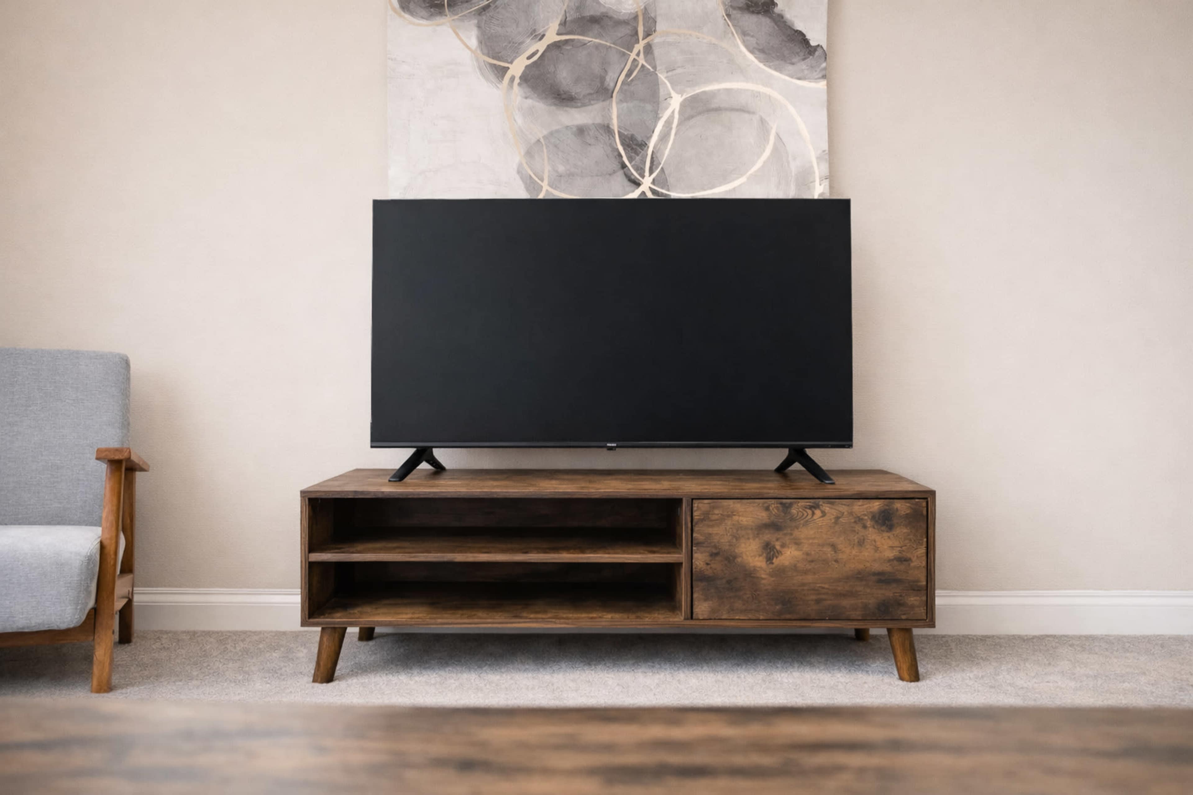 A modern living room features a large black television on a wooden TV stand, accompanied by a simple armchair and a decorative wall art above.