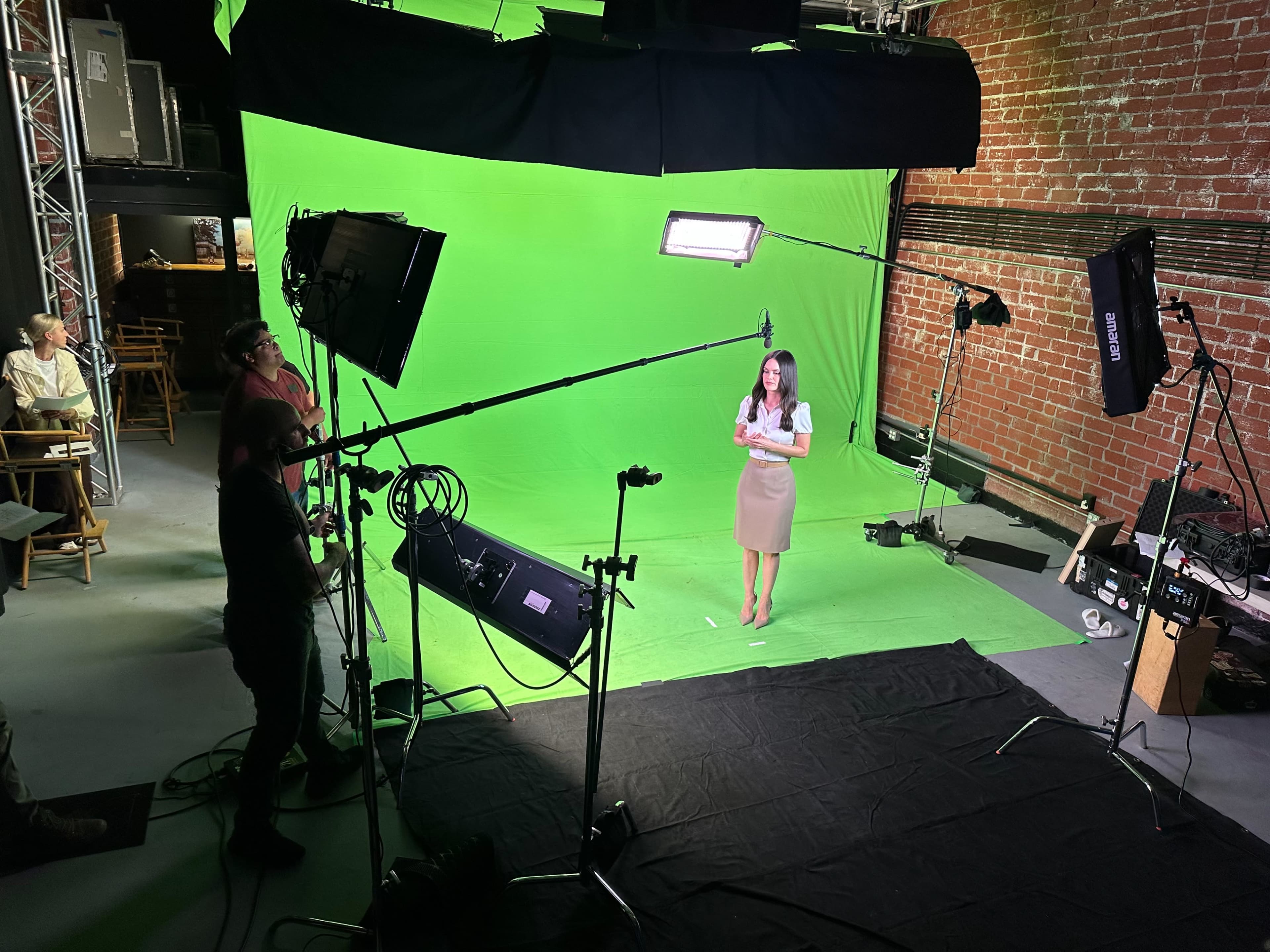 A woman stands on a stage with a green backdrop while crew members operate lighting and cameras around her.