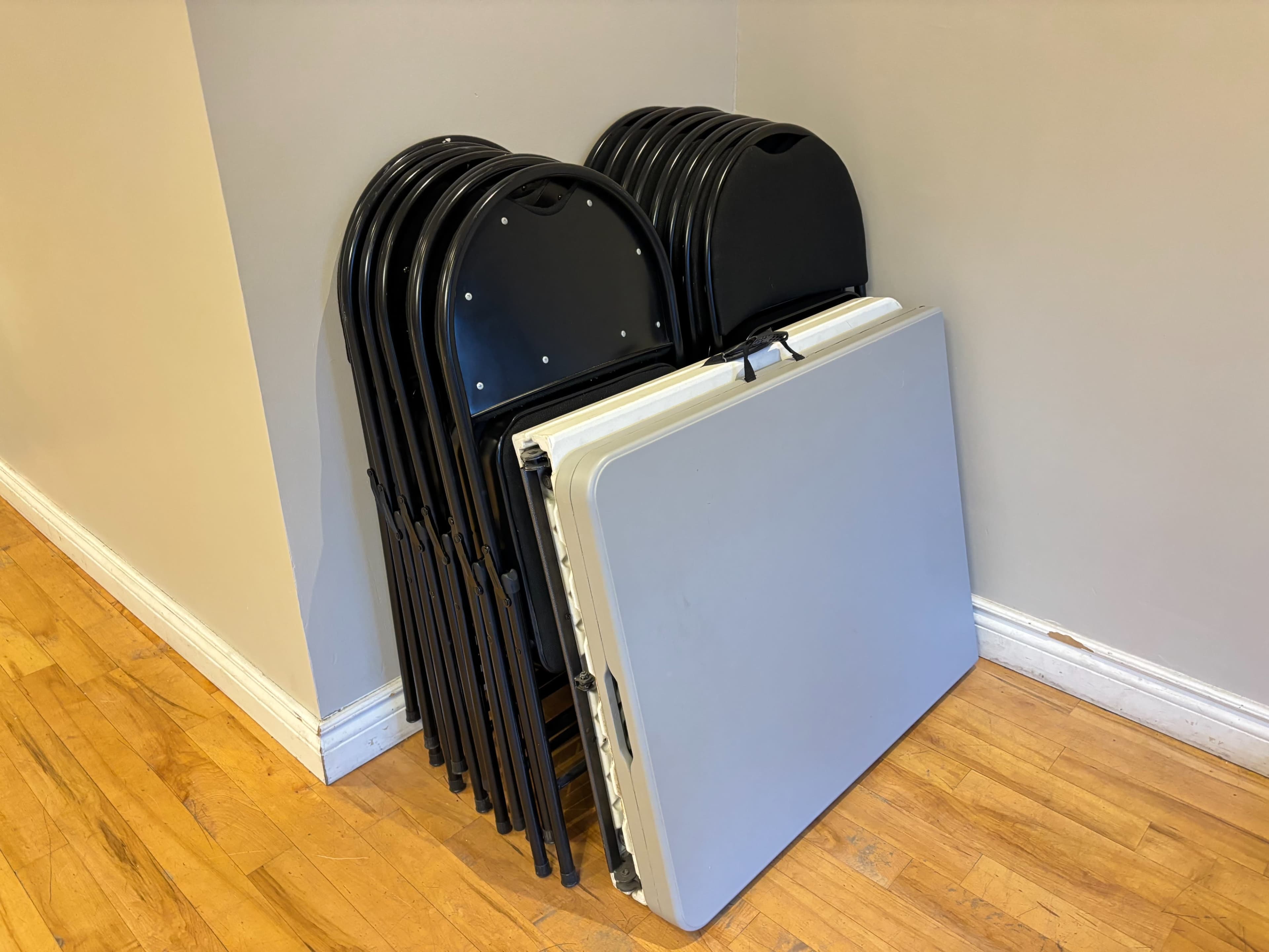 A stack of black folding chairs is positioned next to a folded gray table against a beige wall.
