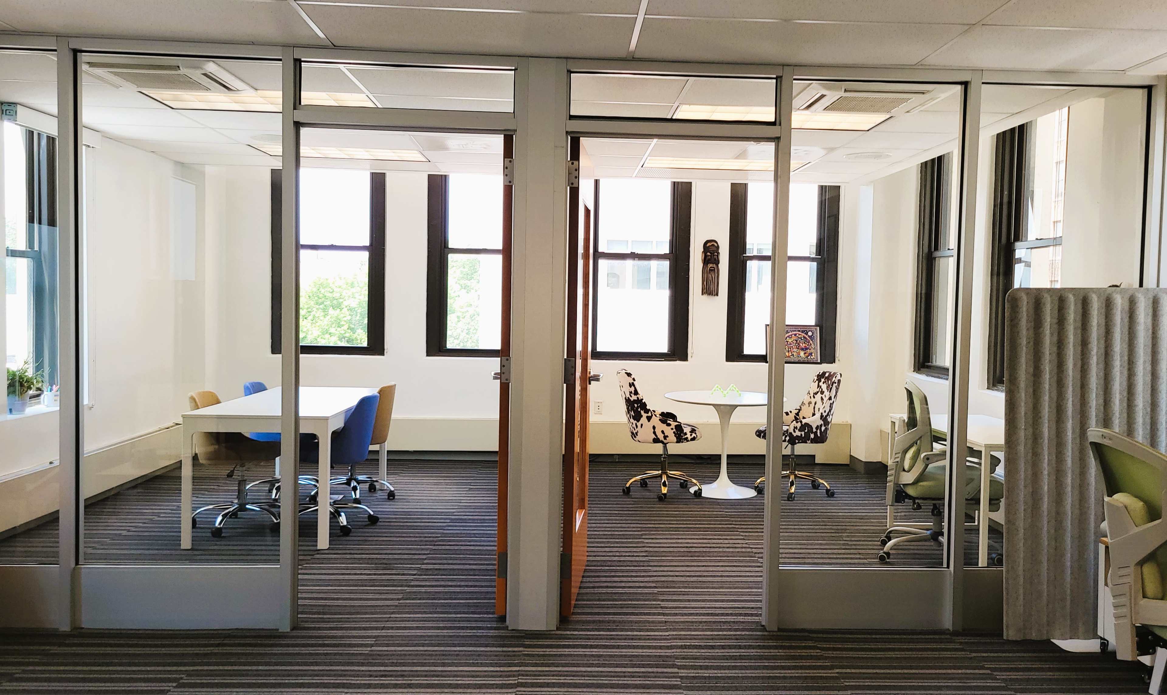 A modern office space with glass walls dividing two meeting rooms, each furnished with tables and chairs.
