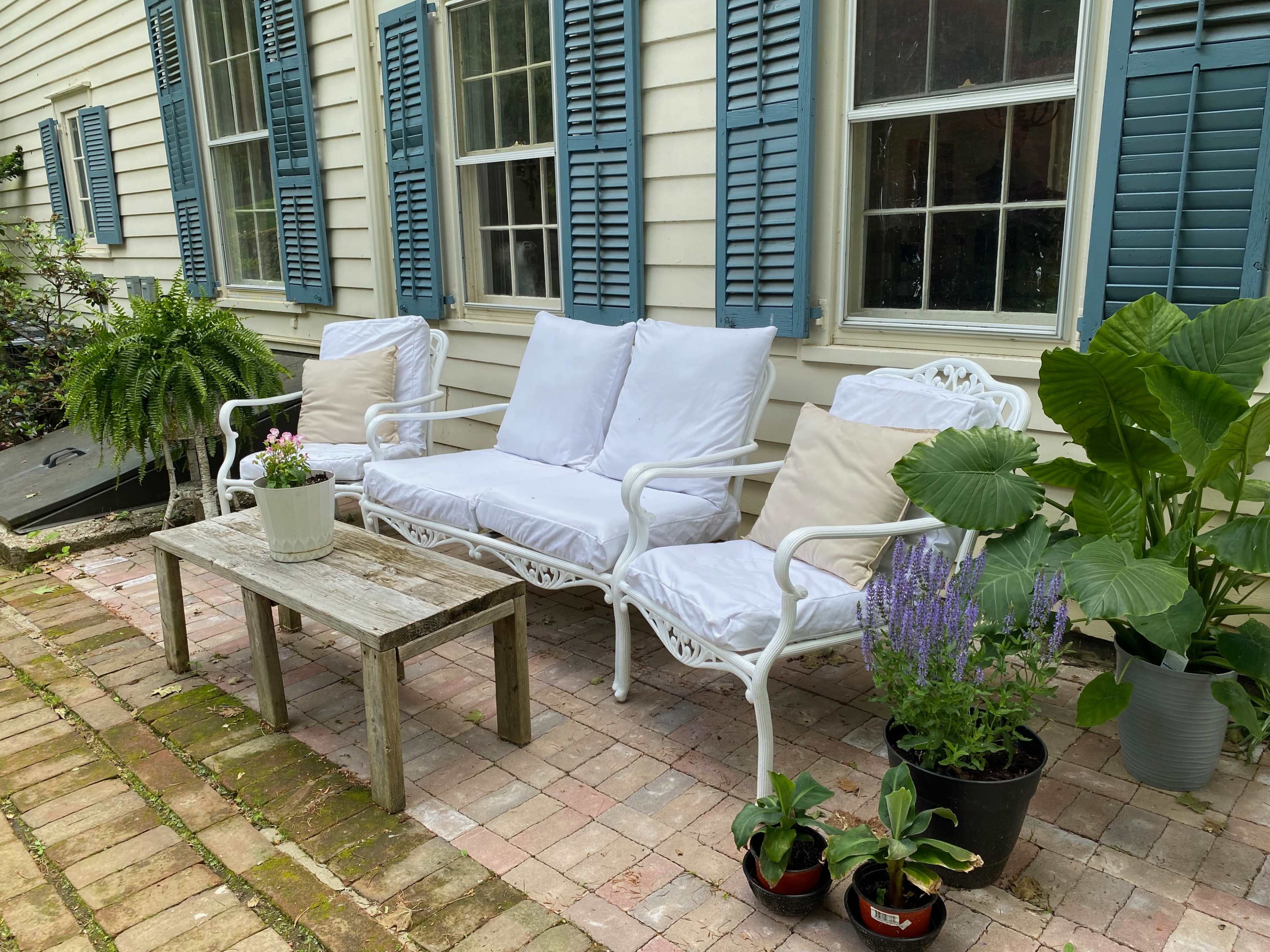 A small outdoor seating area features two white metal chairs with cushions and a wooden table, surrounded by potted plants on a brick patio.