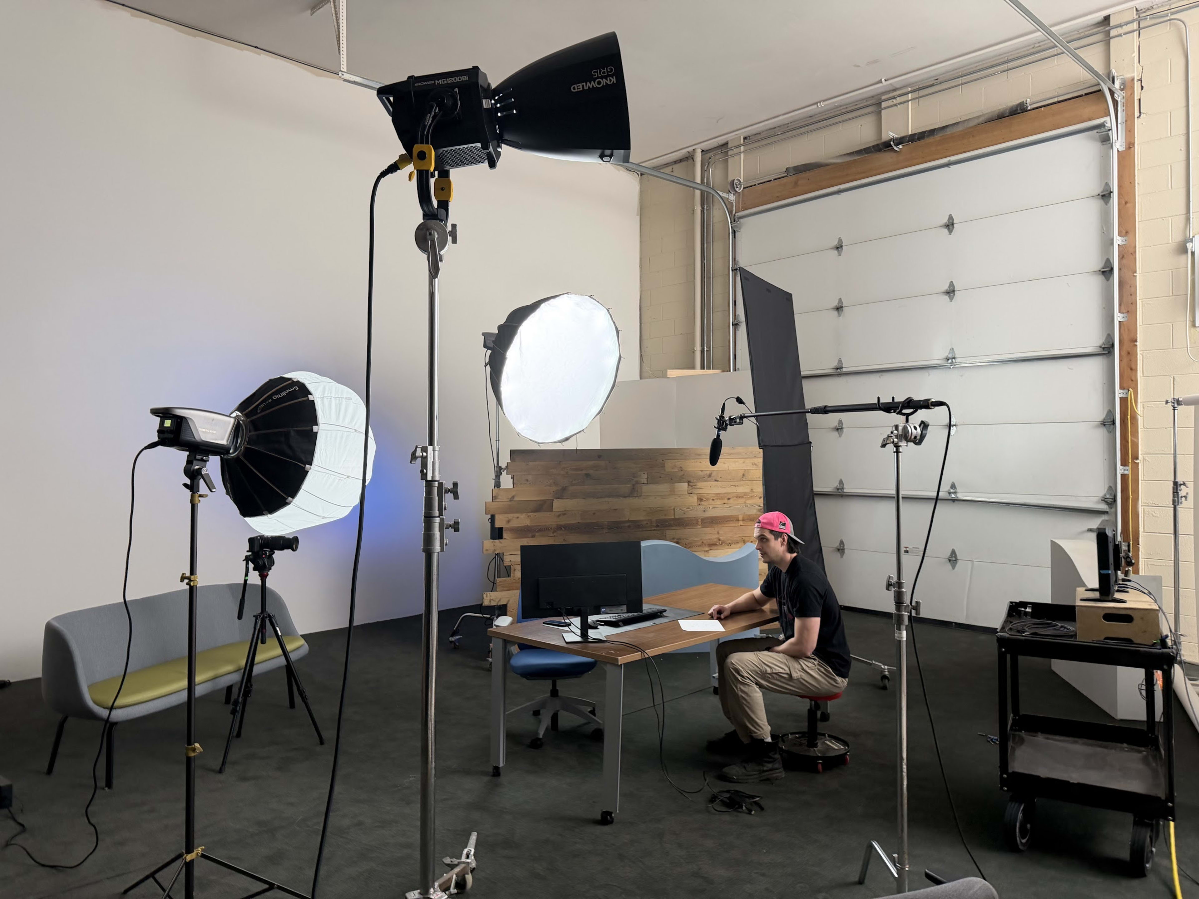 A person is seated at a desk in a studio setup, surrounded by various lighting equipment and a large garage door in the background.