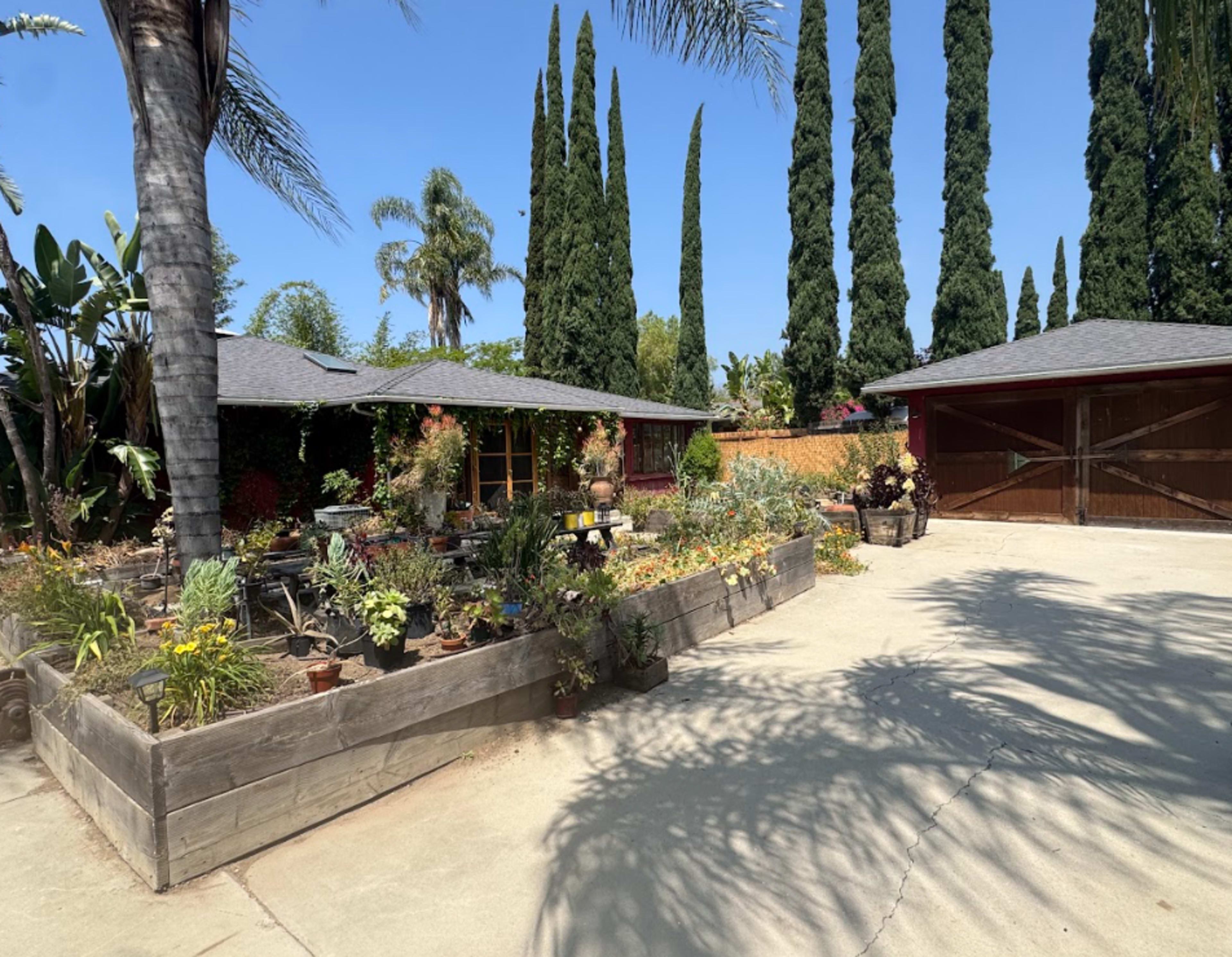 The image shows a landscaped garden with various plants and flowers in raised beds, surrounding a single-story house and a garage with wooden doors.
