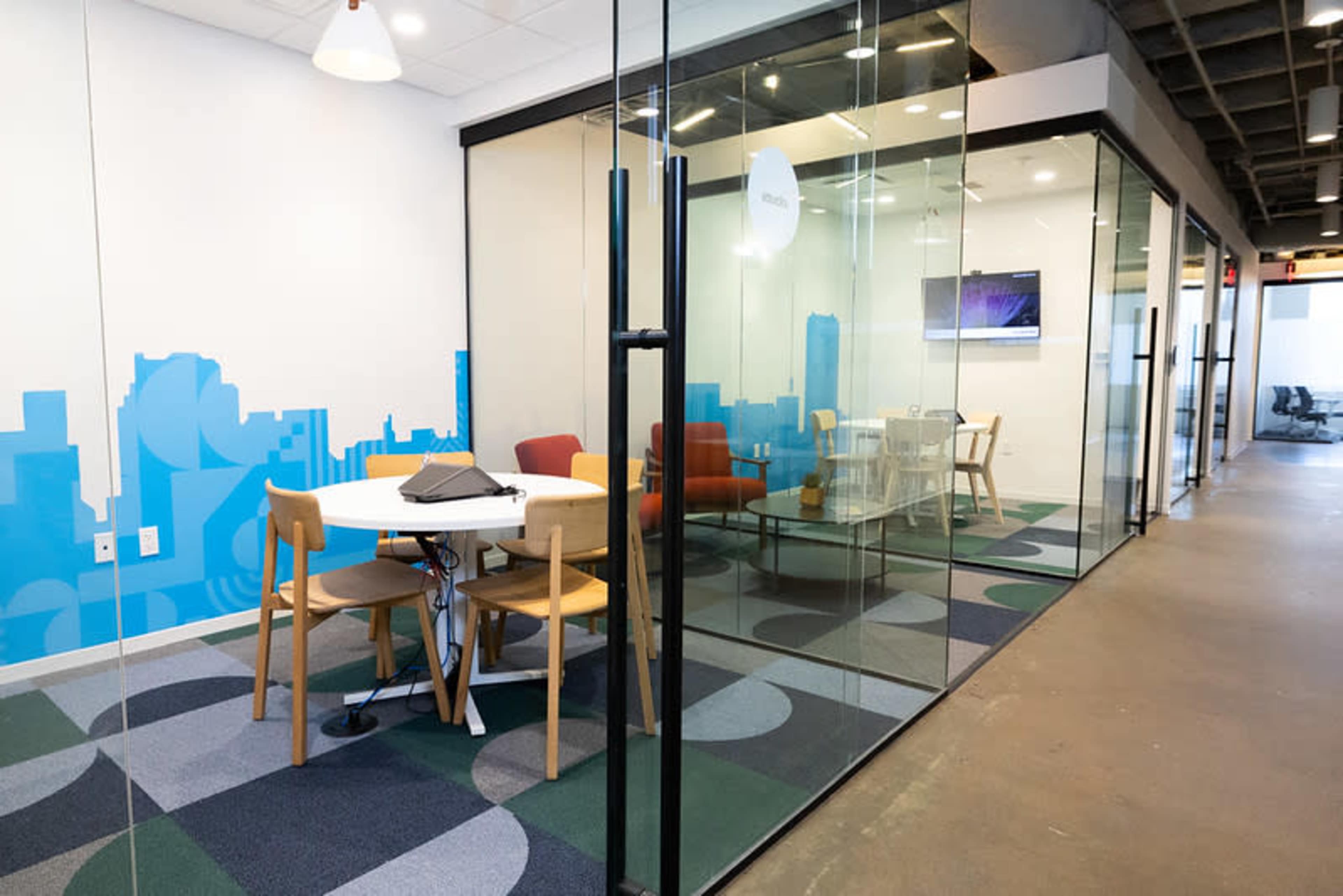 The image shows a modern office space with glass-walled meeting rooms, a round table with chairs, and a lounge area featuring a sofa.