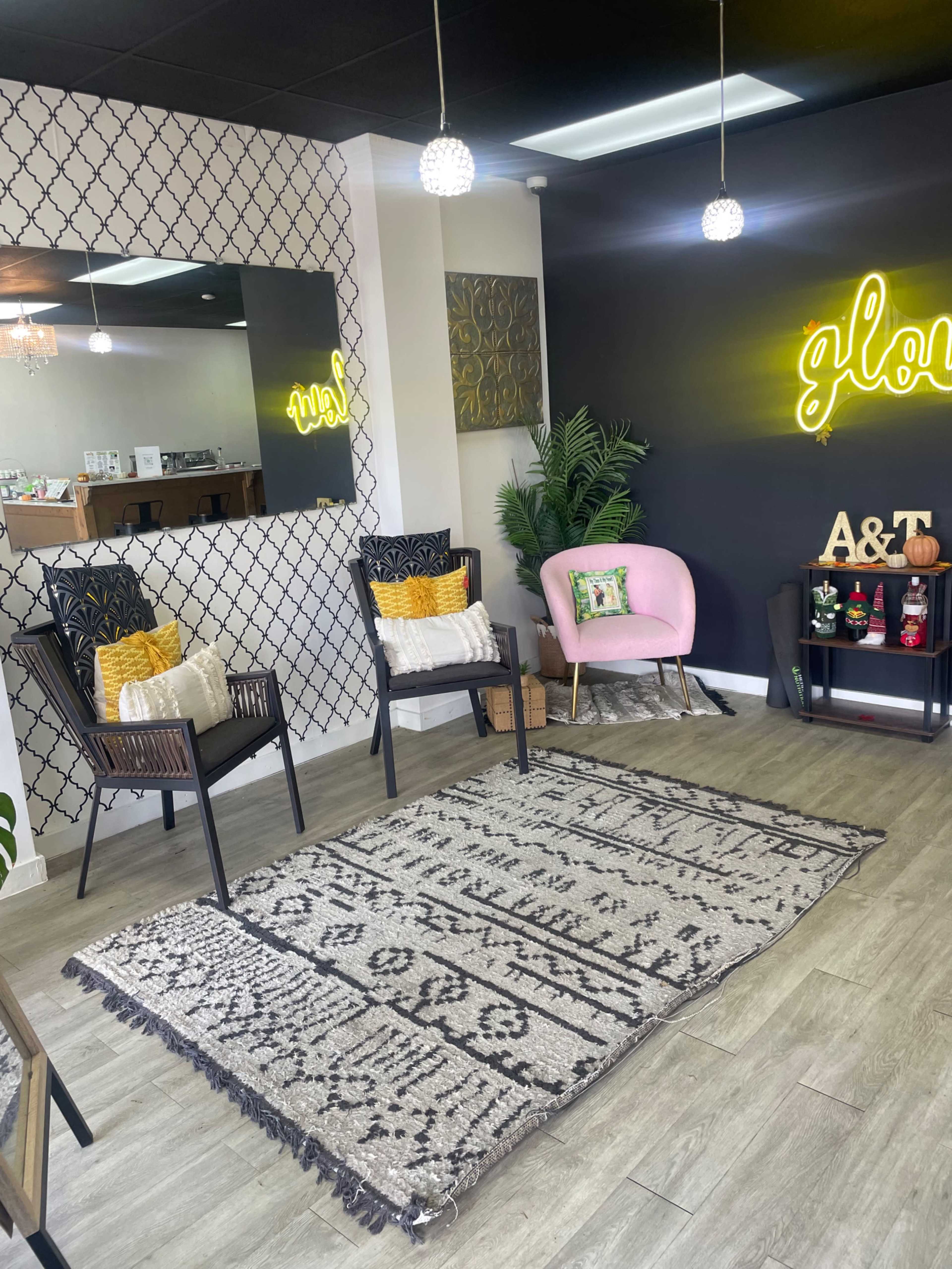The image shows a cozy waiting area with two chairs, a pink accent chair, a patterned rug, and a neon sign on the wall.