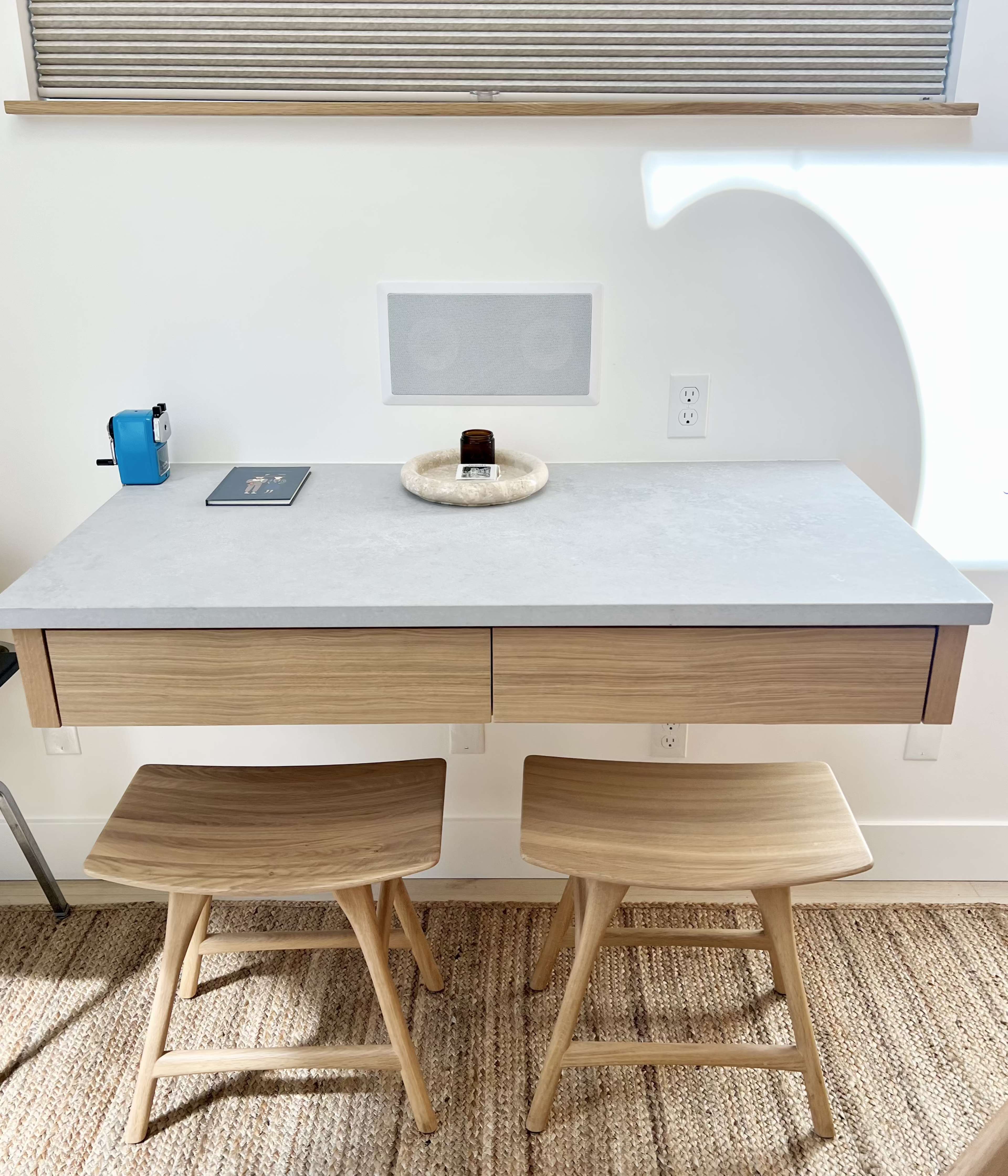 A wooden desk with two chairs and a small decorative dish sits against a white wall with a speaker and a window above it.