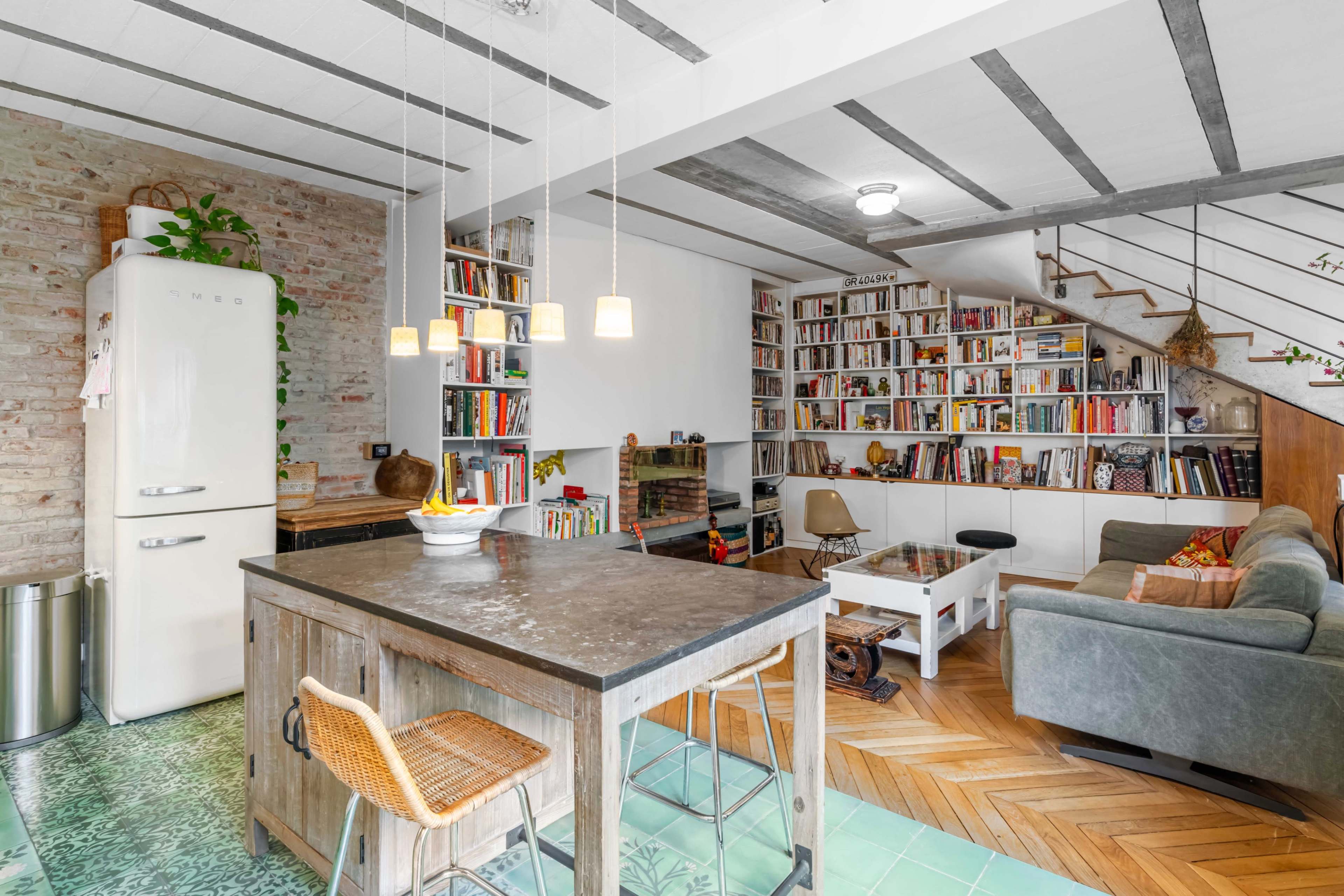 A modern kitchen and living area with a wooden table, a retro refrigerator, bookshelves filled with books, and a staircase leading to an upper level.