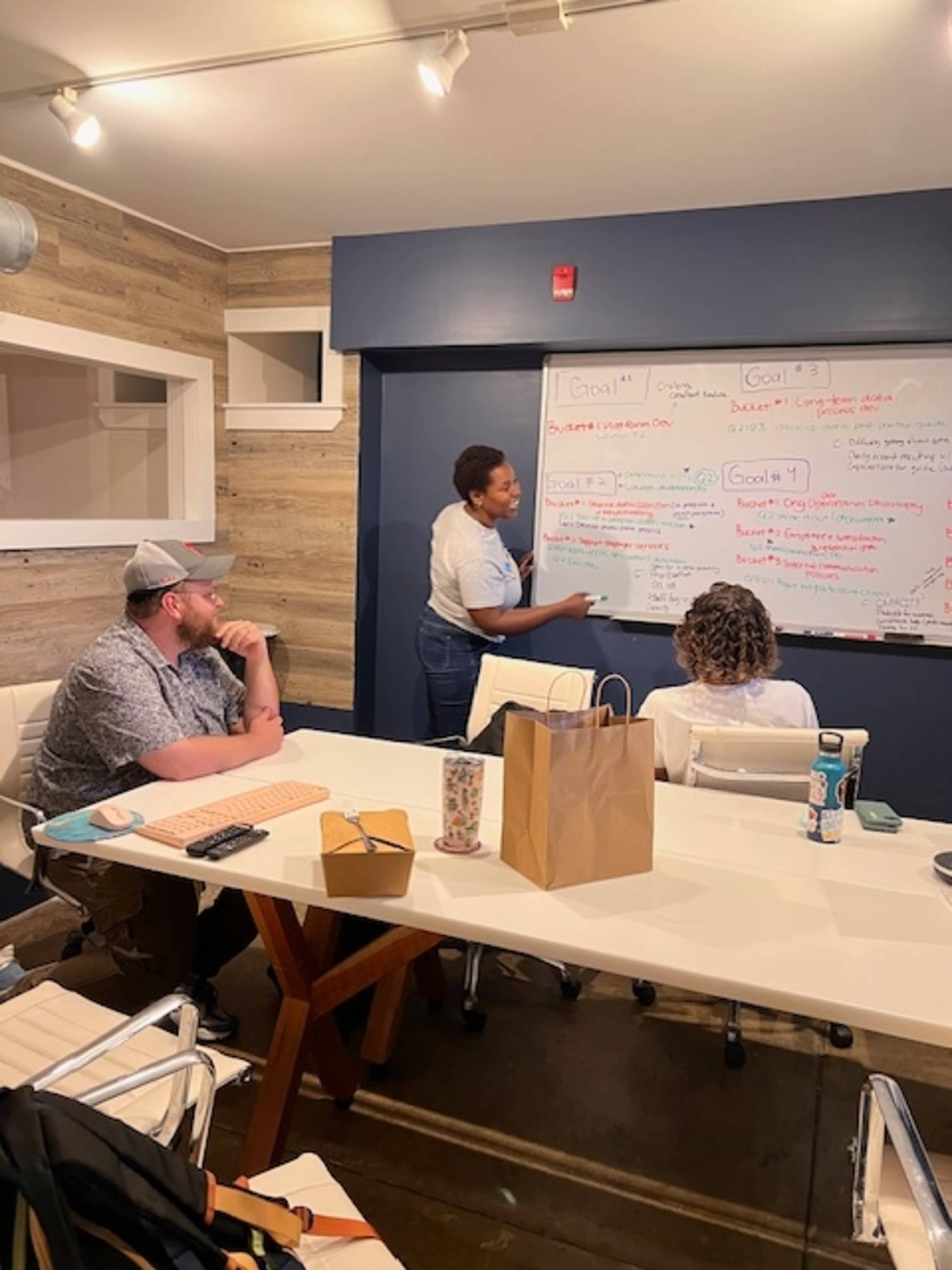 A person is presenting at a whiteboard while two others sit at a table in a modern meeting room.