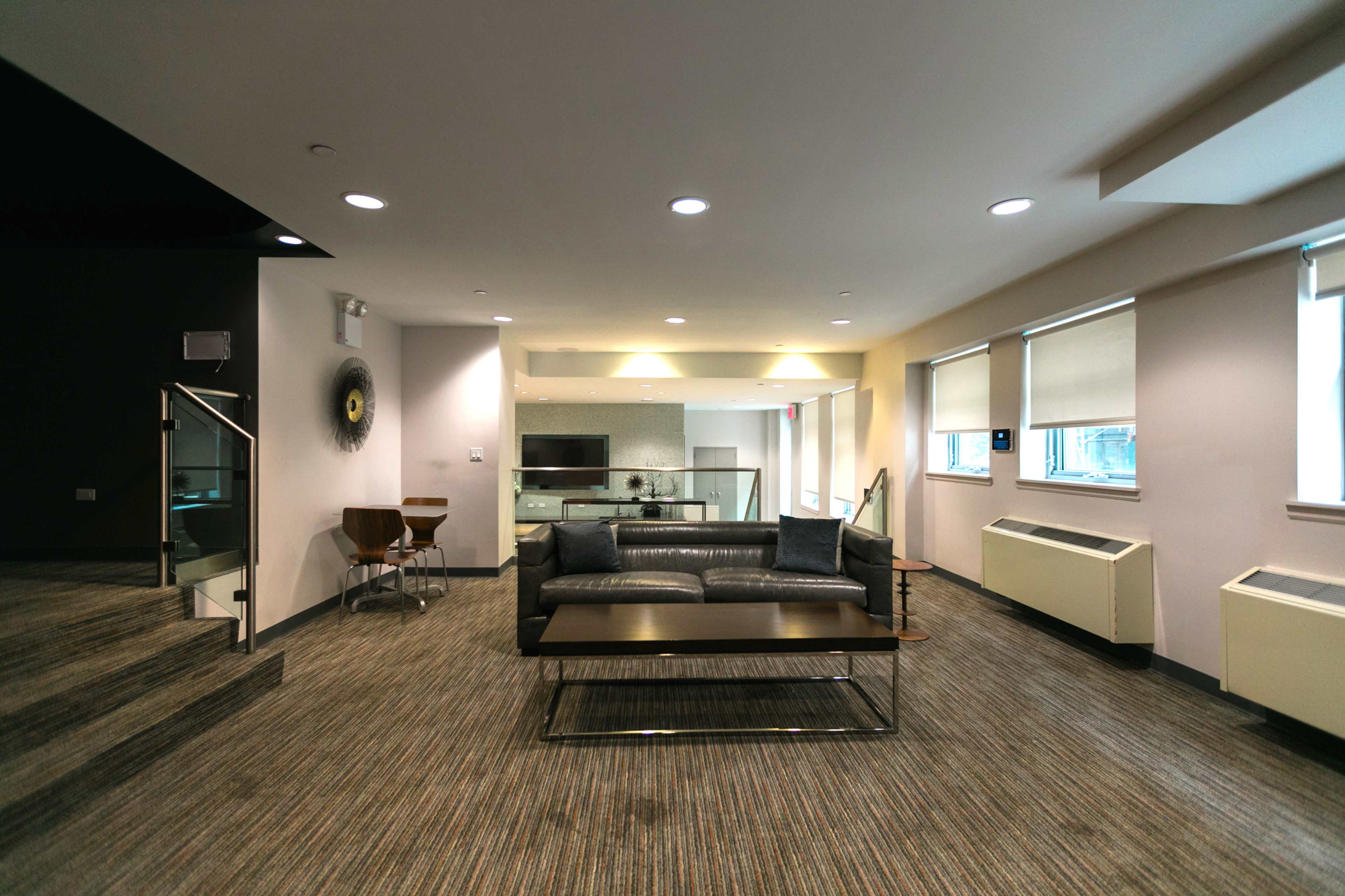 The image shows a modern lounge area featuring a gray sofa, a coffee table, and large windows, with a staircase leading to an upper level.