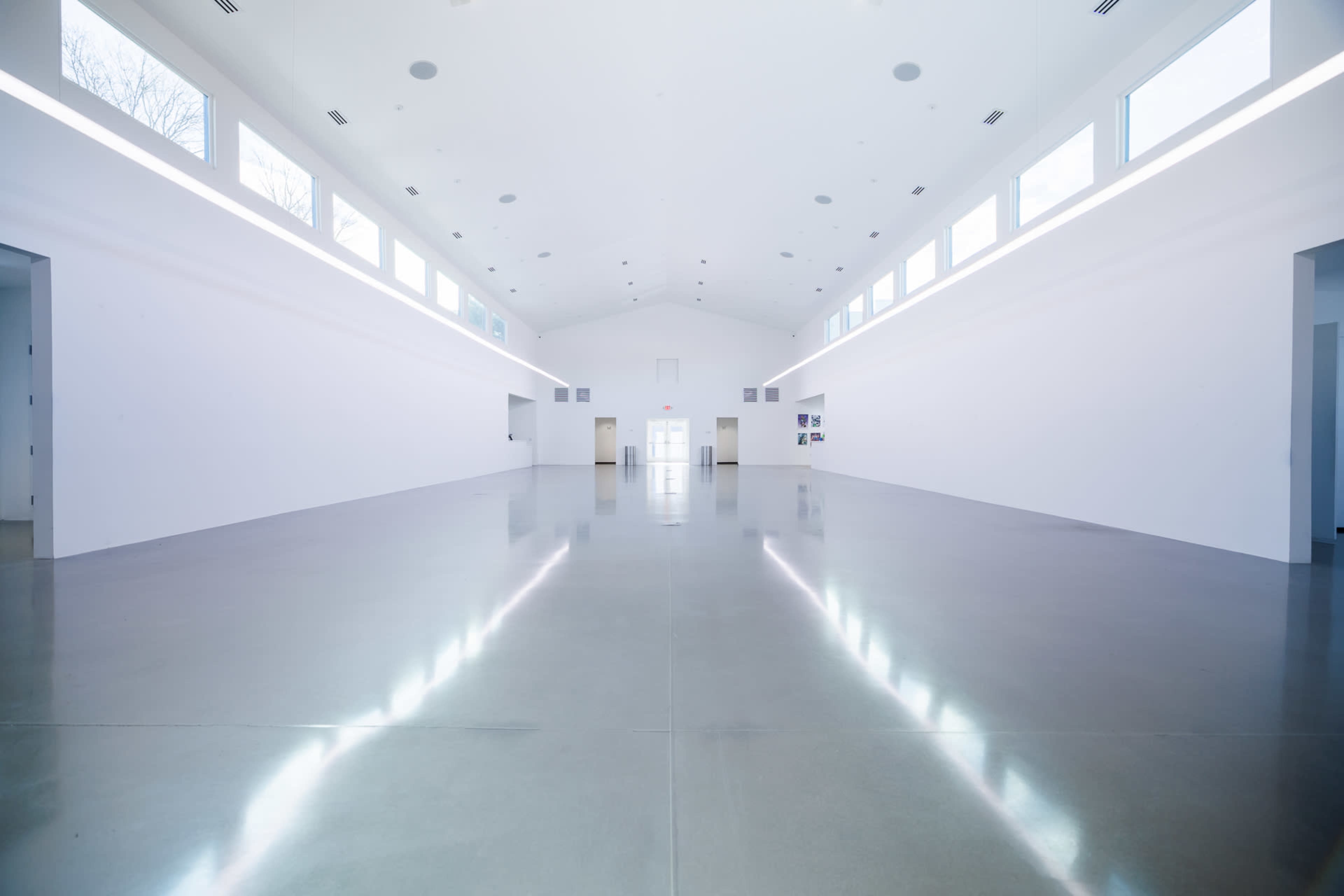 The image depicts a spacious, minimalistic interior with white walls, concrete flooring, and large windows allowing light to flood the area.