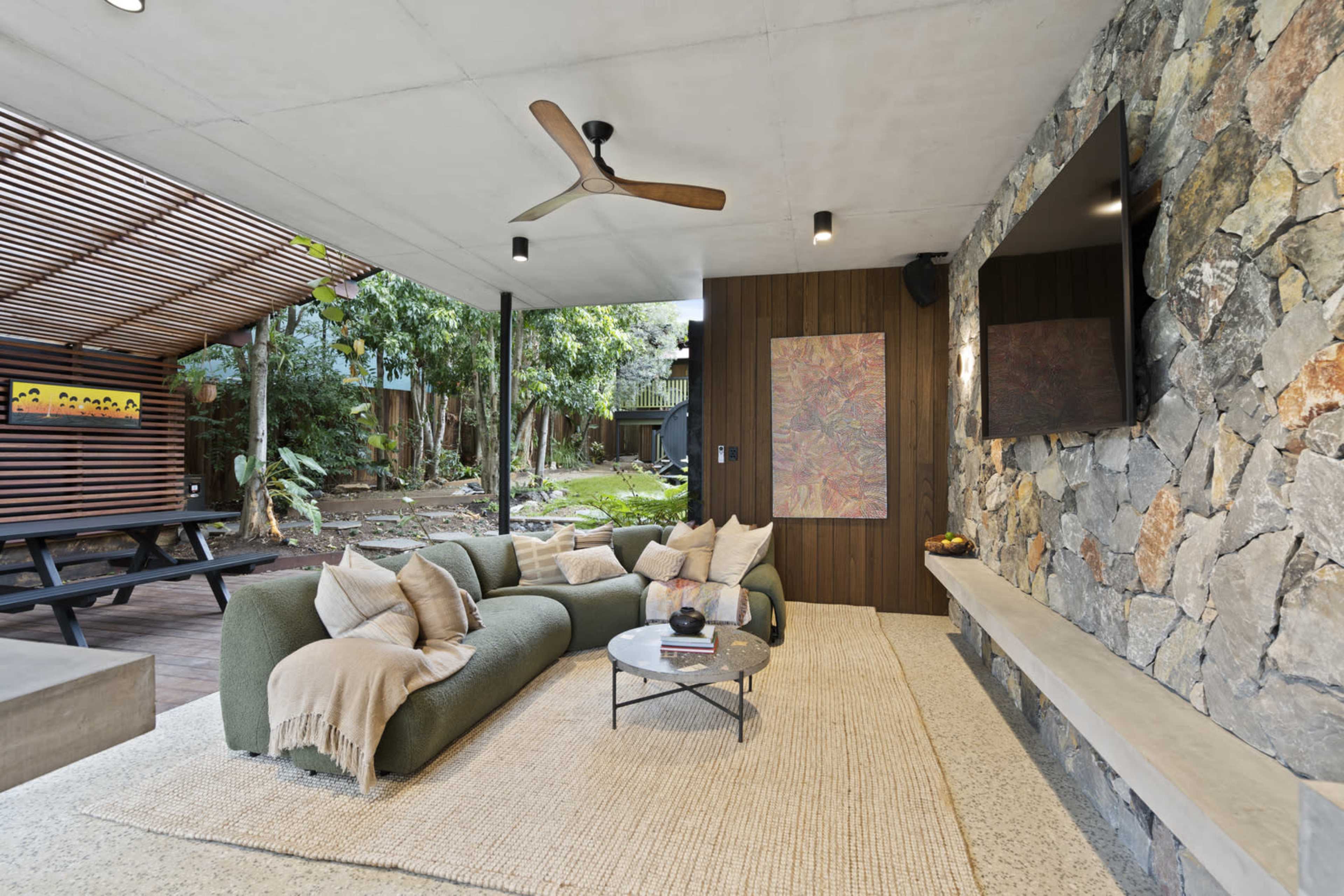 The image depicts a modern outdoor living space featuring a stone accent wall, comfortable seating, and a coffee table, surrounded by lush greenery.