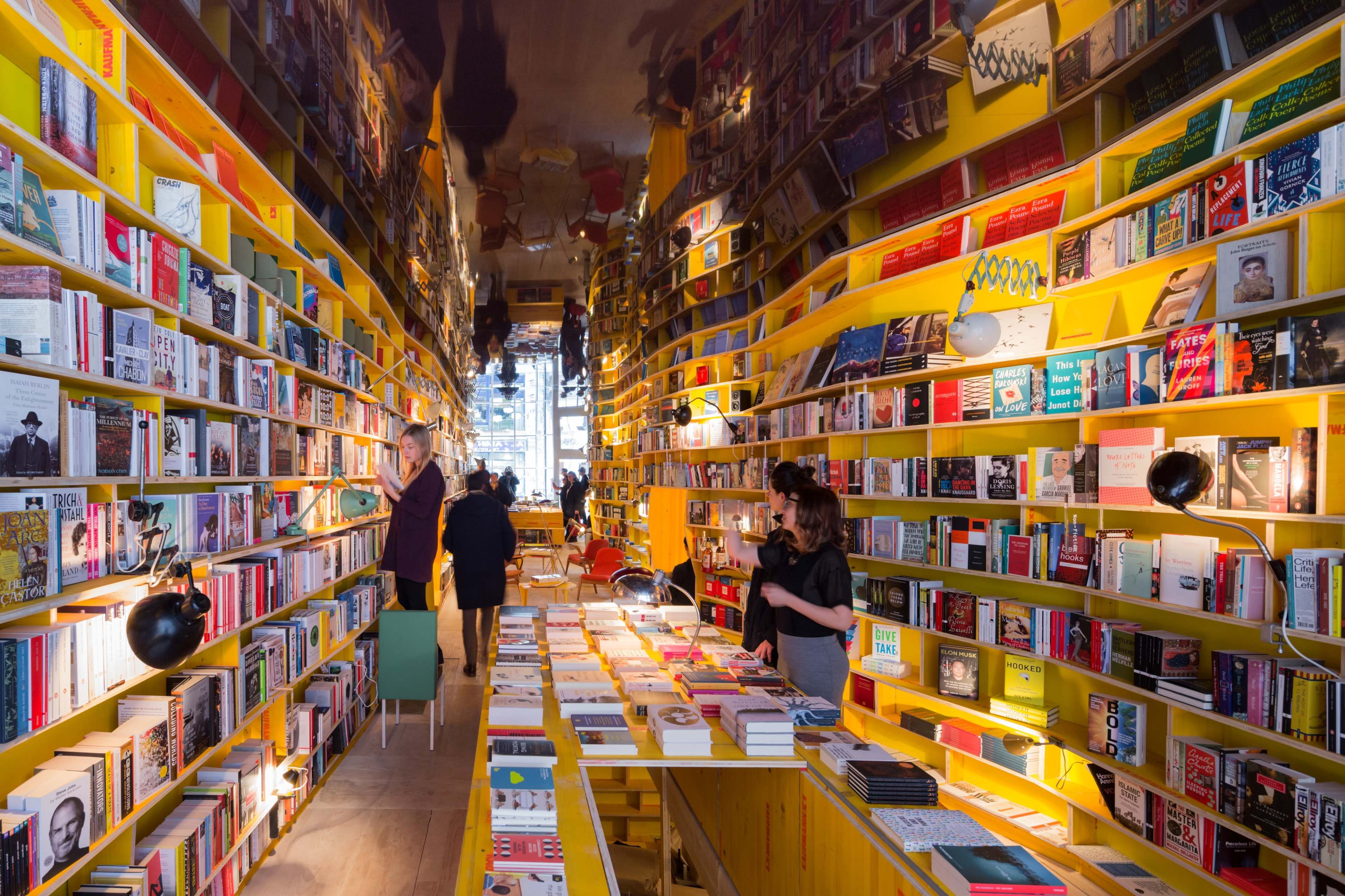 The image shows a brightly lit bookstore with walls lined with colorful bookshelves and people browsing among tables filled with books.
