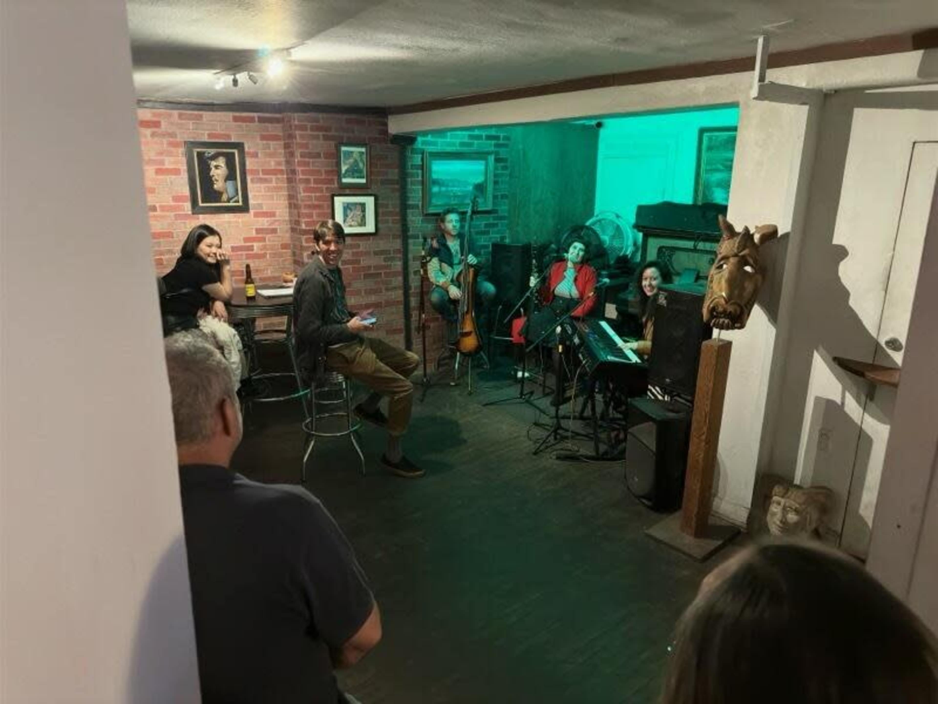 A small music performance is taking place in a cozy room with a few seated audience members and musicians playing instruments.
