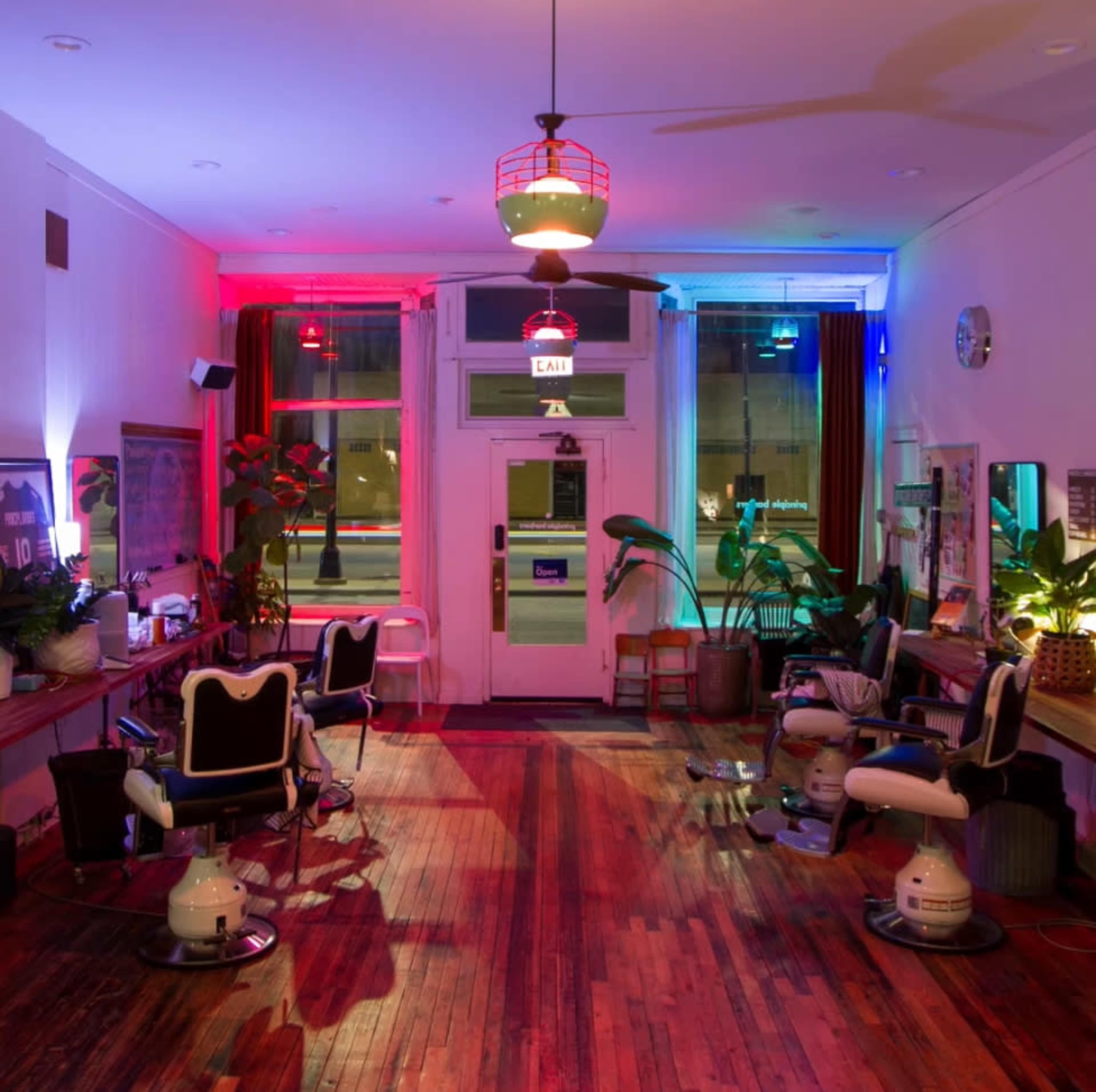A brightly lit barber shop features vintage-style barber chairs and plants, with colorful lighting illuminating the wooden floor and walls.