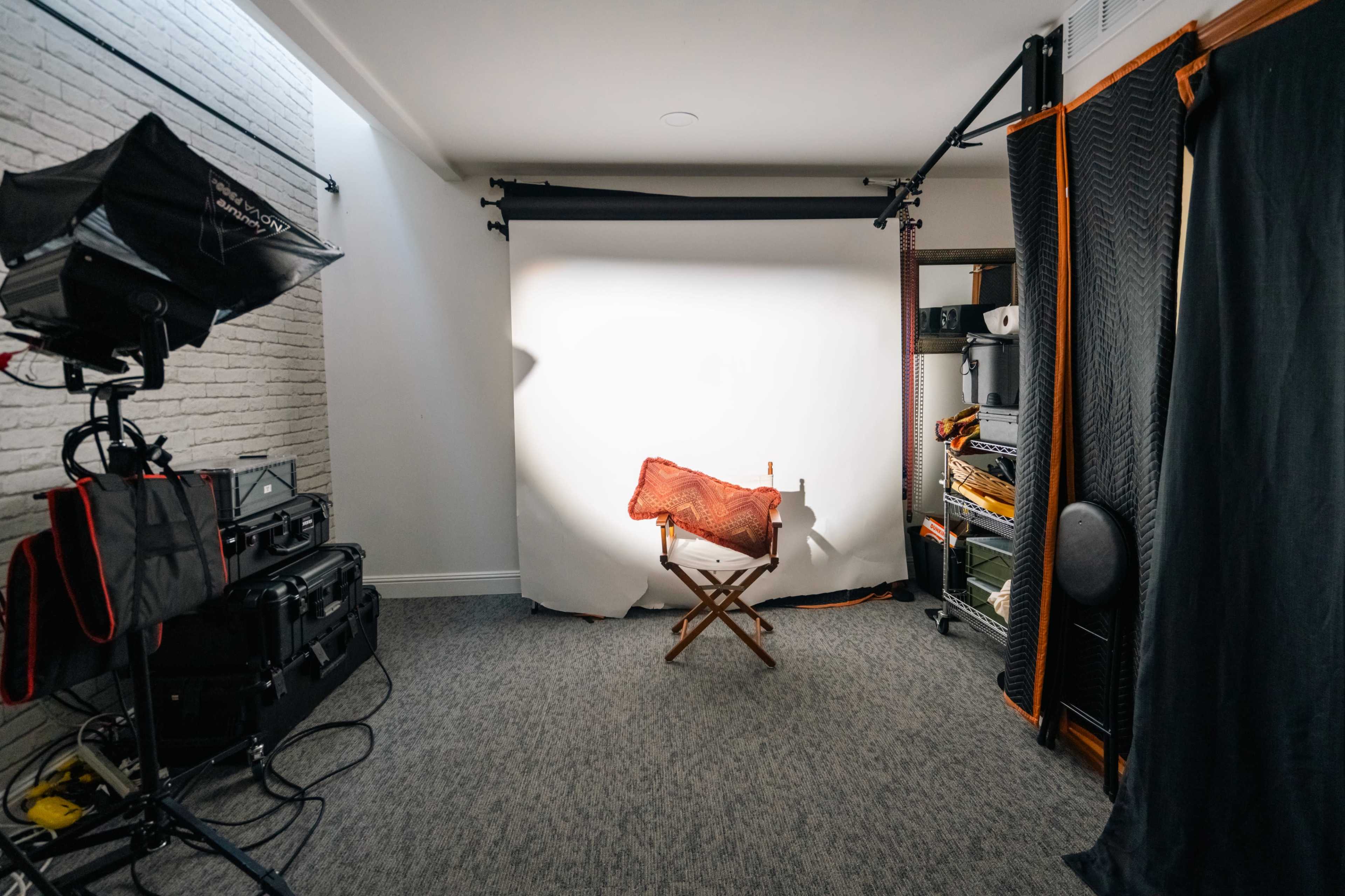 A photography studio with a blank white backdrop, a director's chair, and various lighting equipment arranged around the space.