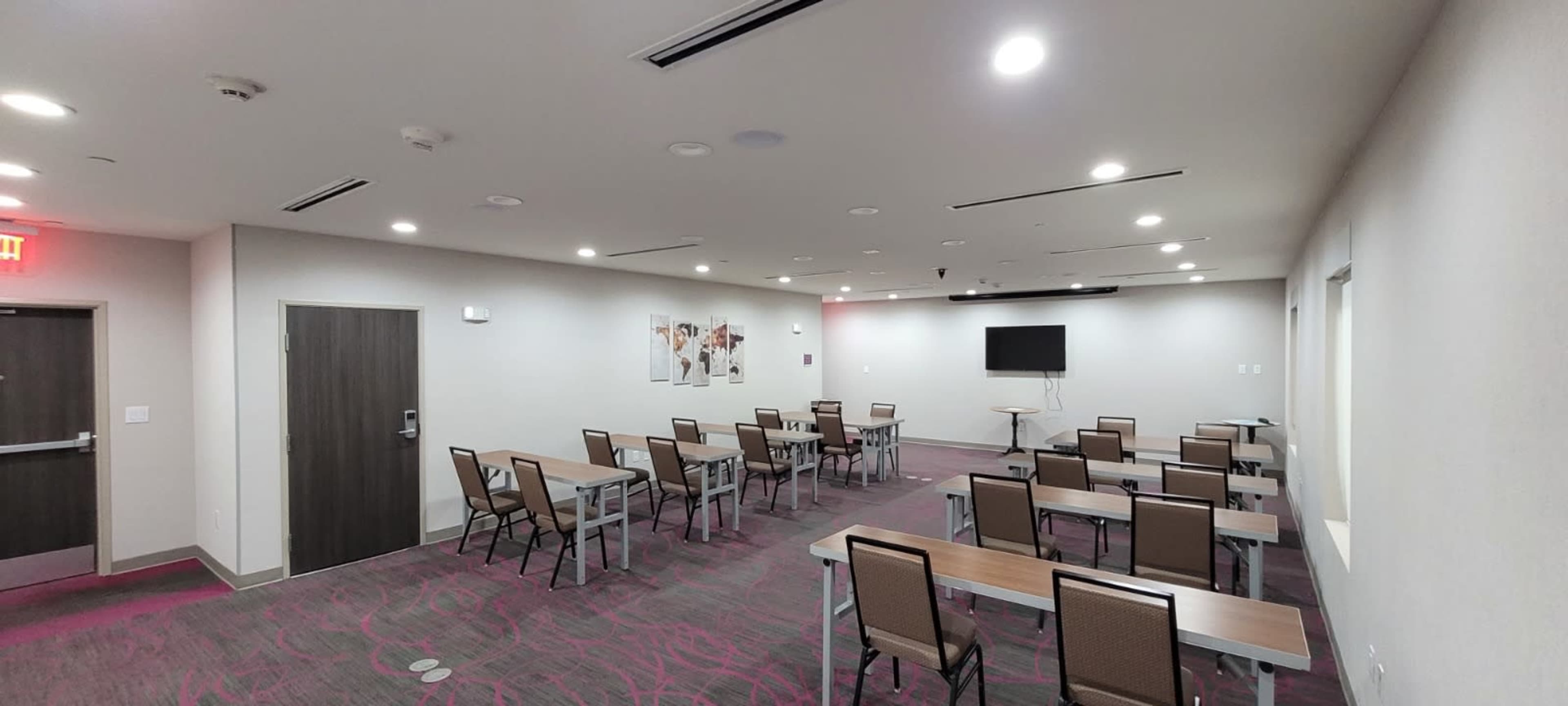 The image shows a conference room with several rectangular tables arranged in rows and a wall-mounted screen at the front.