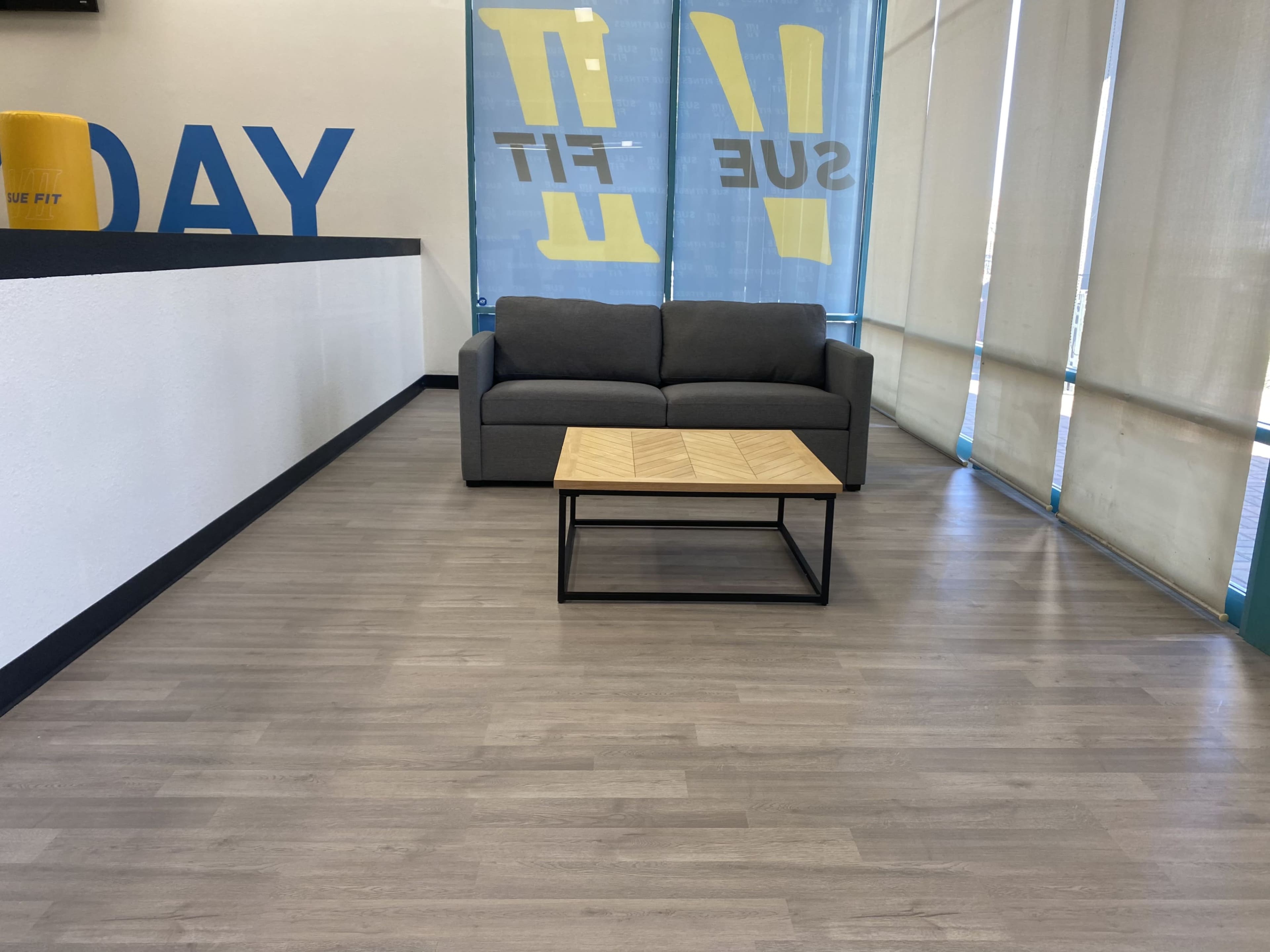 A gray sofa is positioned in front of a wooden coffee table in a brightly lit room with large windows and motivational banners on the walls.