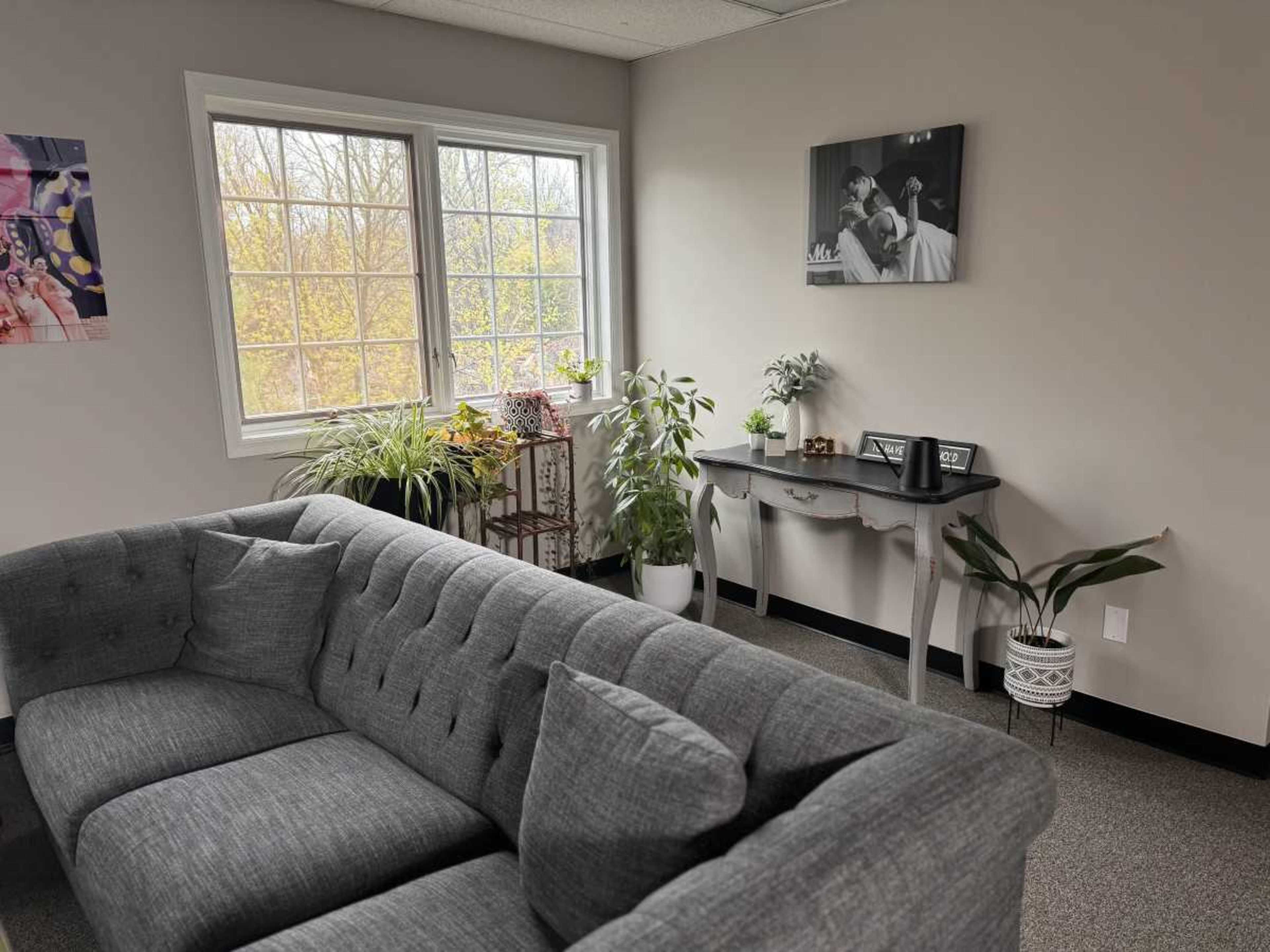 The image shows a corner of a cozy office space featuring a gray tufted couch, a small table with decorative plants, and large windows with a view of trees.