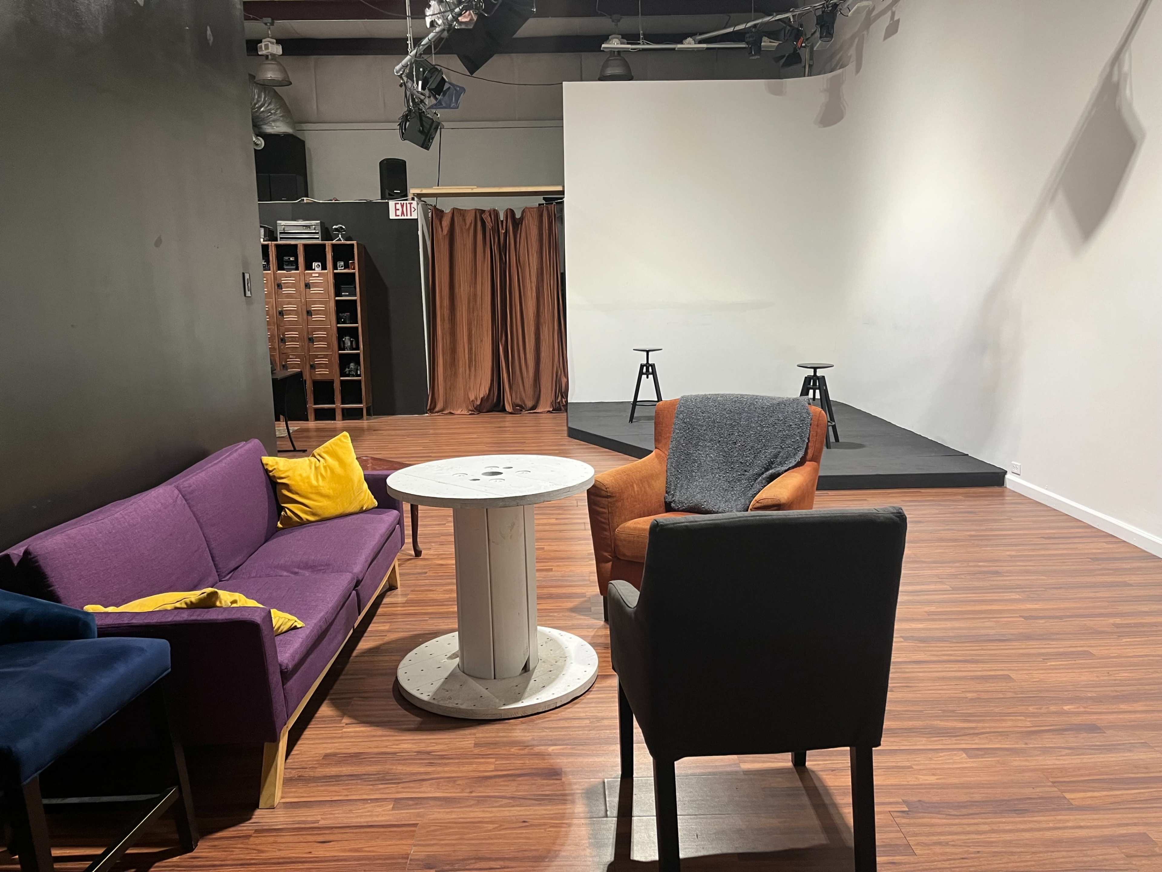 A small performance space featuring a seating area with colorful couches and a circular table, facing a small stage with two stools.