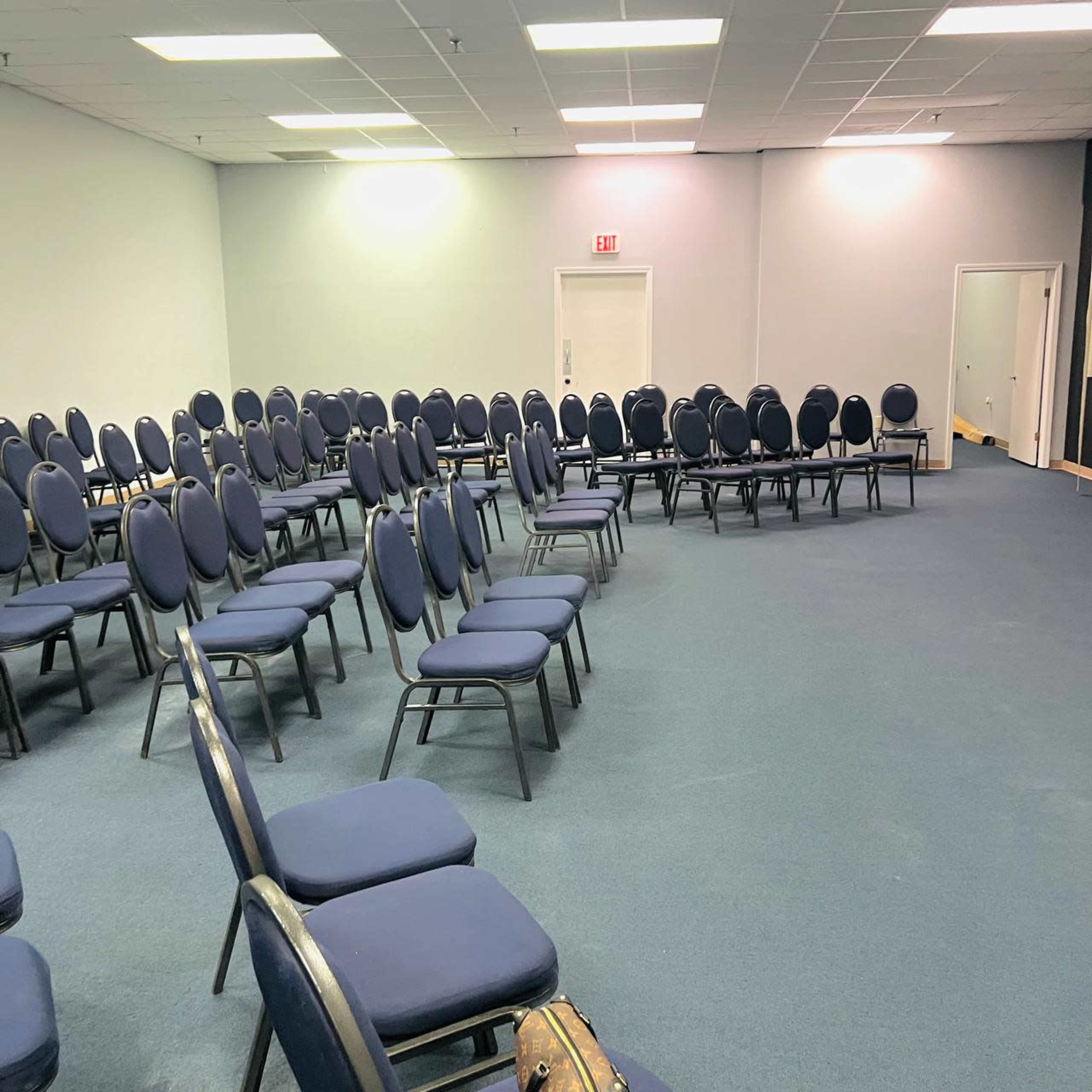 A room with several arranged rows of blue chairs on a carpeted floor, with a door and a small area in the background.