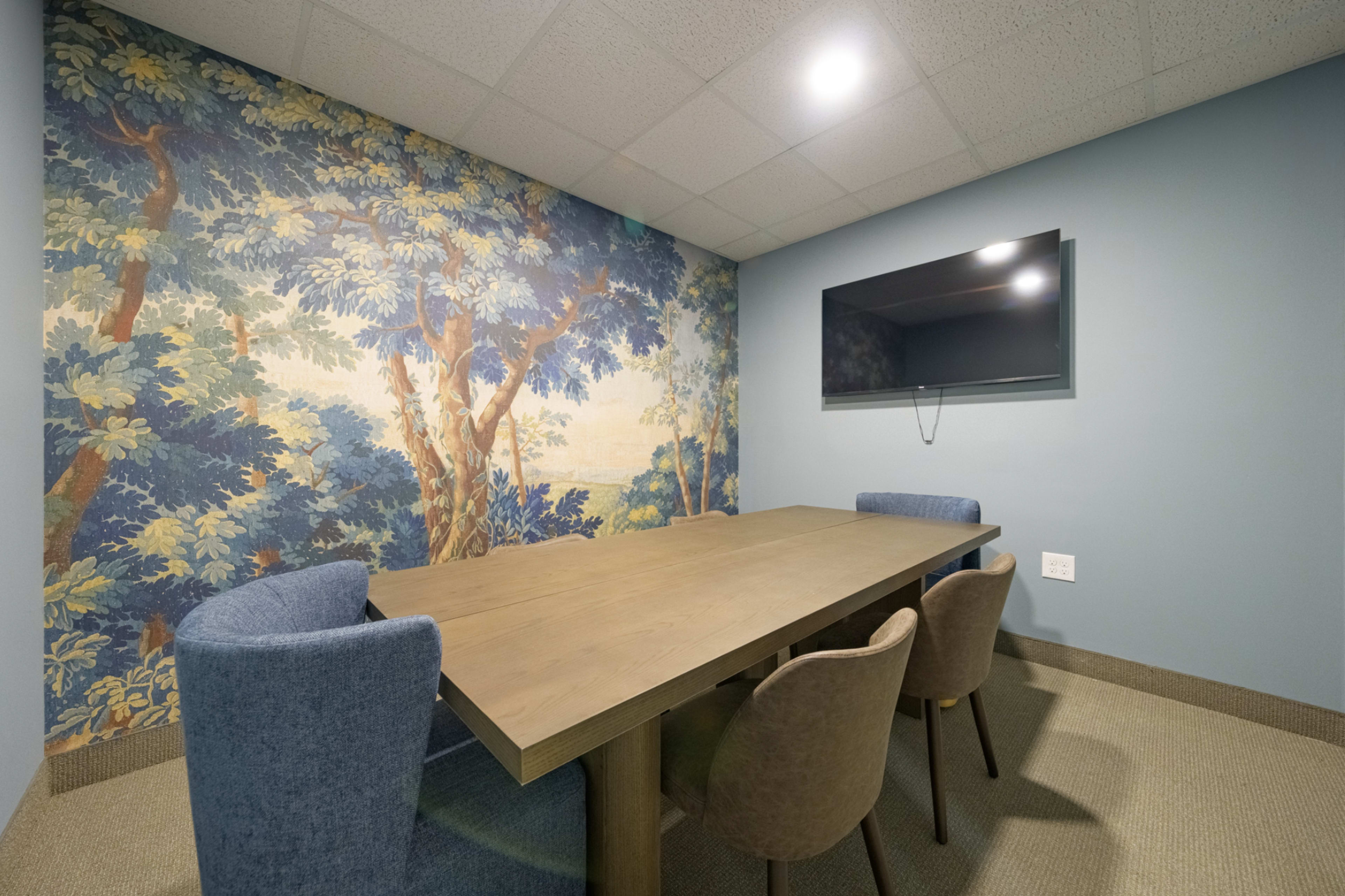 The image shows a small meeting room featuring a wooden table surrounded by four chairs, with a large television mounted on a wall adorned with a mural of trees.
