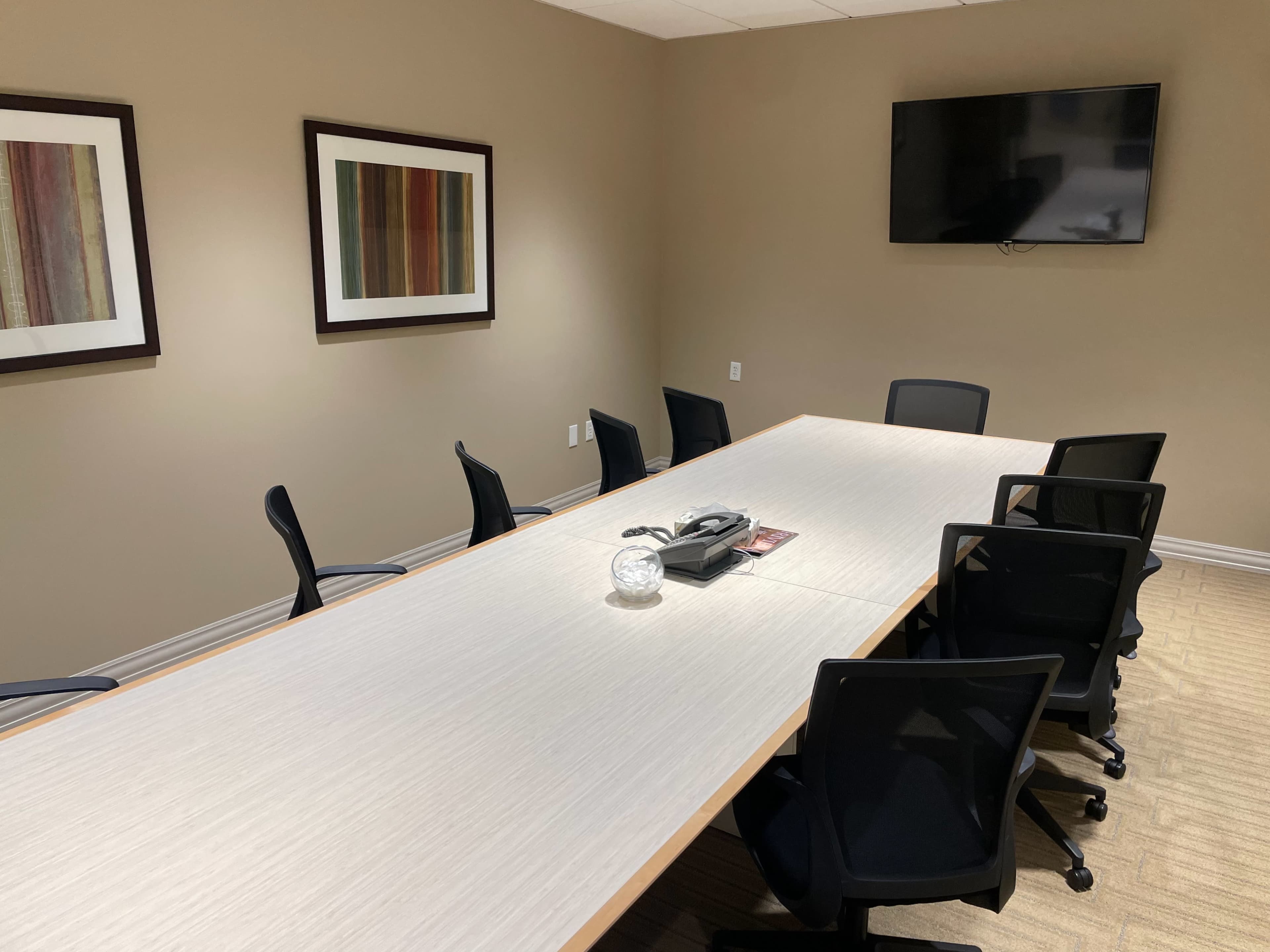 The image shows a conference room with a long wooden table surrounded by black chairs, a phone, and a wall-mounted television along with abstract artwork on the walls.