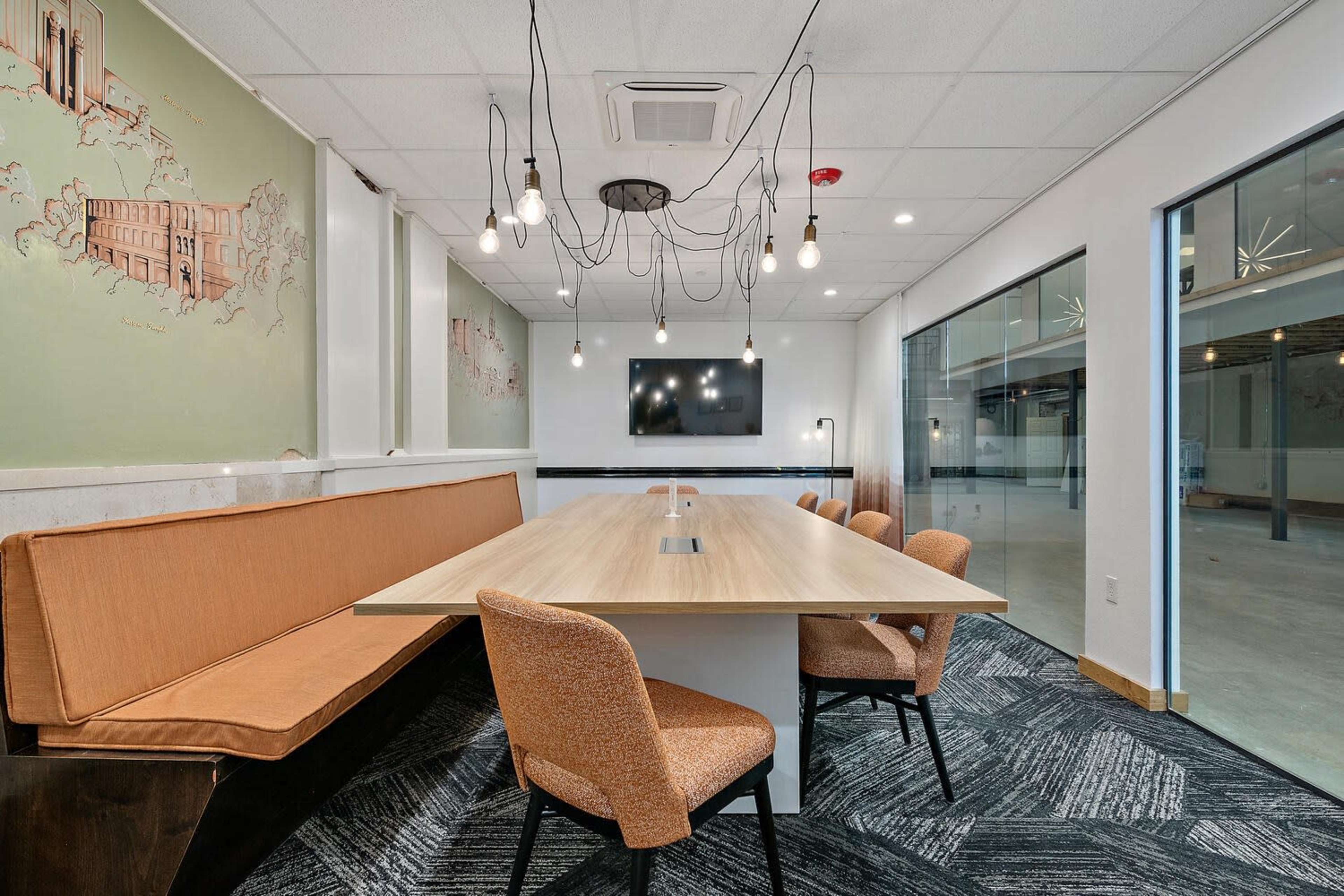 The image shows a modern conference room featuring a long wooden table surrounded by orange chairs, and decorative wall murals in a well-lit space.