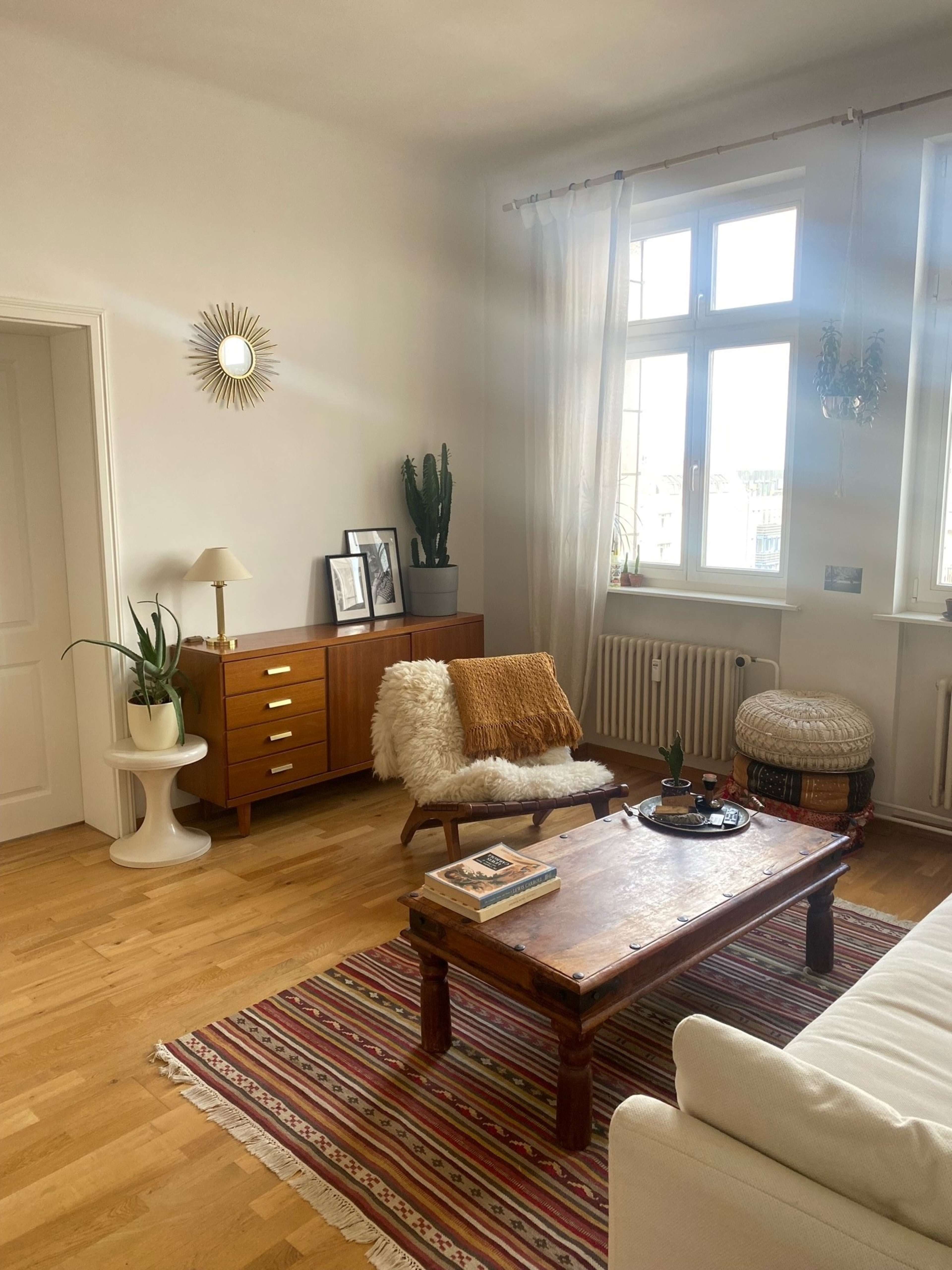 A cozy living room with a wooden coffee table, a plush armchair, and a sideboard, all set against a backdrop of large windows and a colorful rug.