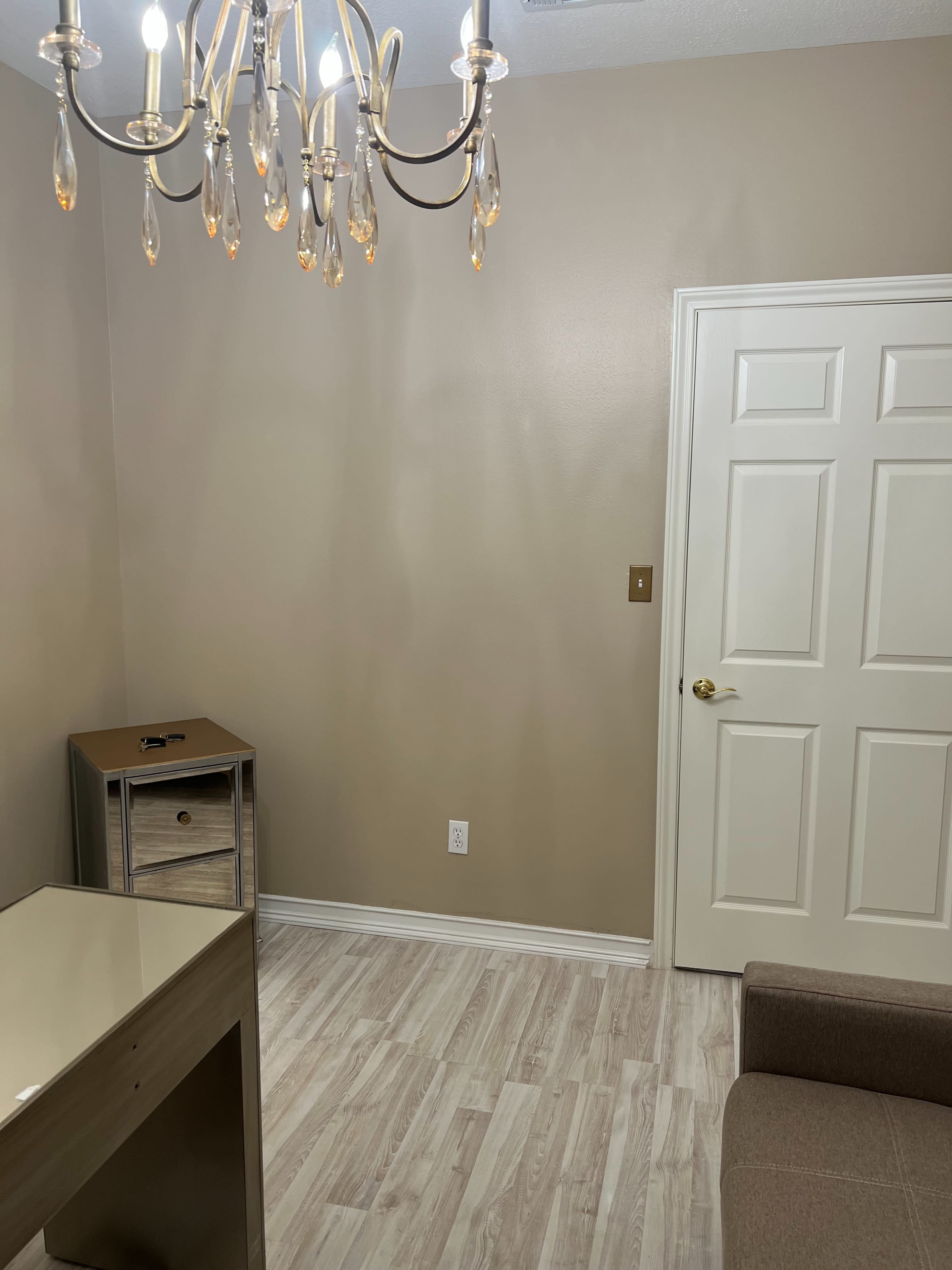 A small room with a chandelier, beige walls, a light-colored floor, a gray couch, a cabinet with a drawer, and a closed white door.