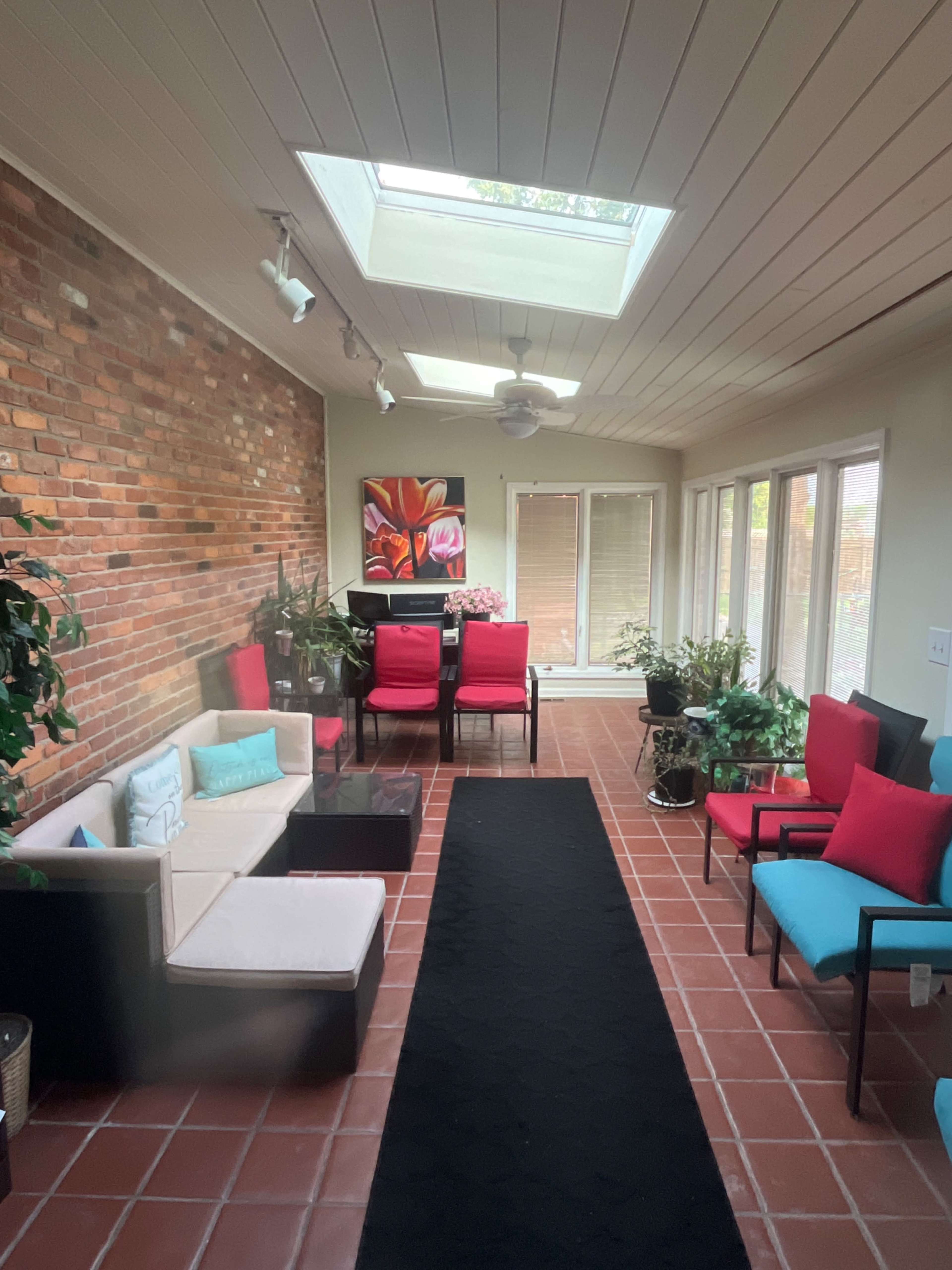 The image shows a sunlit sunroom with a mix of seating options, a large skylight, and brick walls, featuring various plants and colorful cushions.