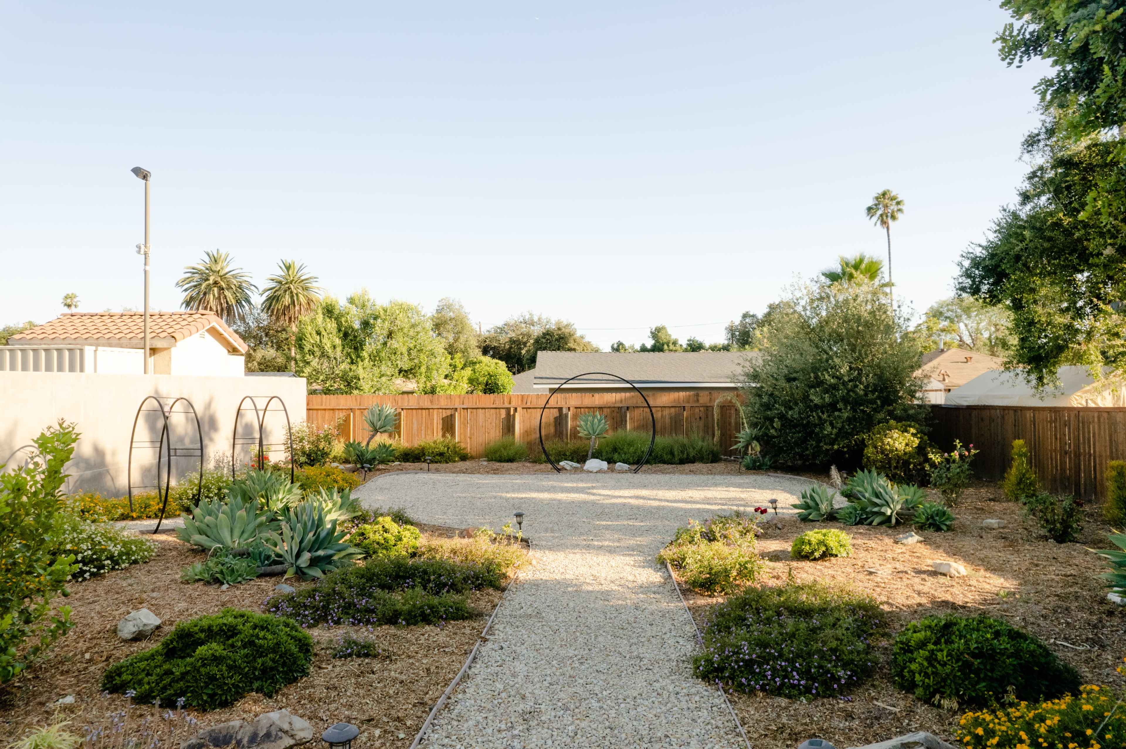 A landscaped backyard featuring gravel pathways, various plants, and two arch structures in a sunny setting.