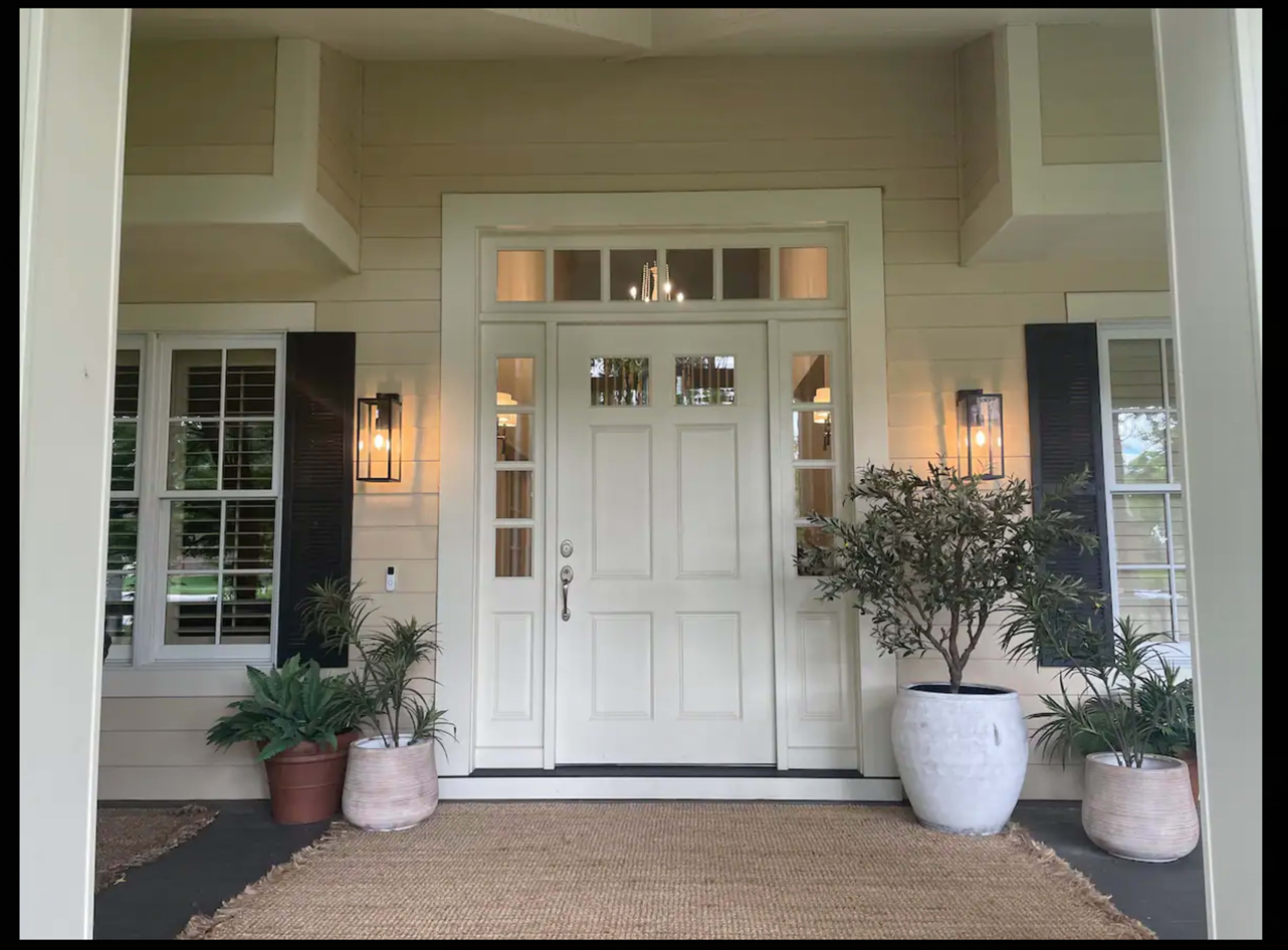A white front door with glass panels is flanked by two plants in pots and is set within a light-colored house with black shutters.