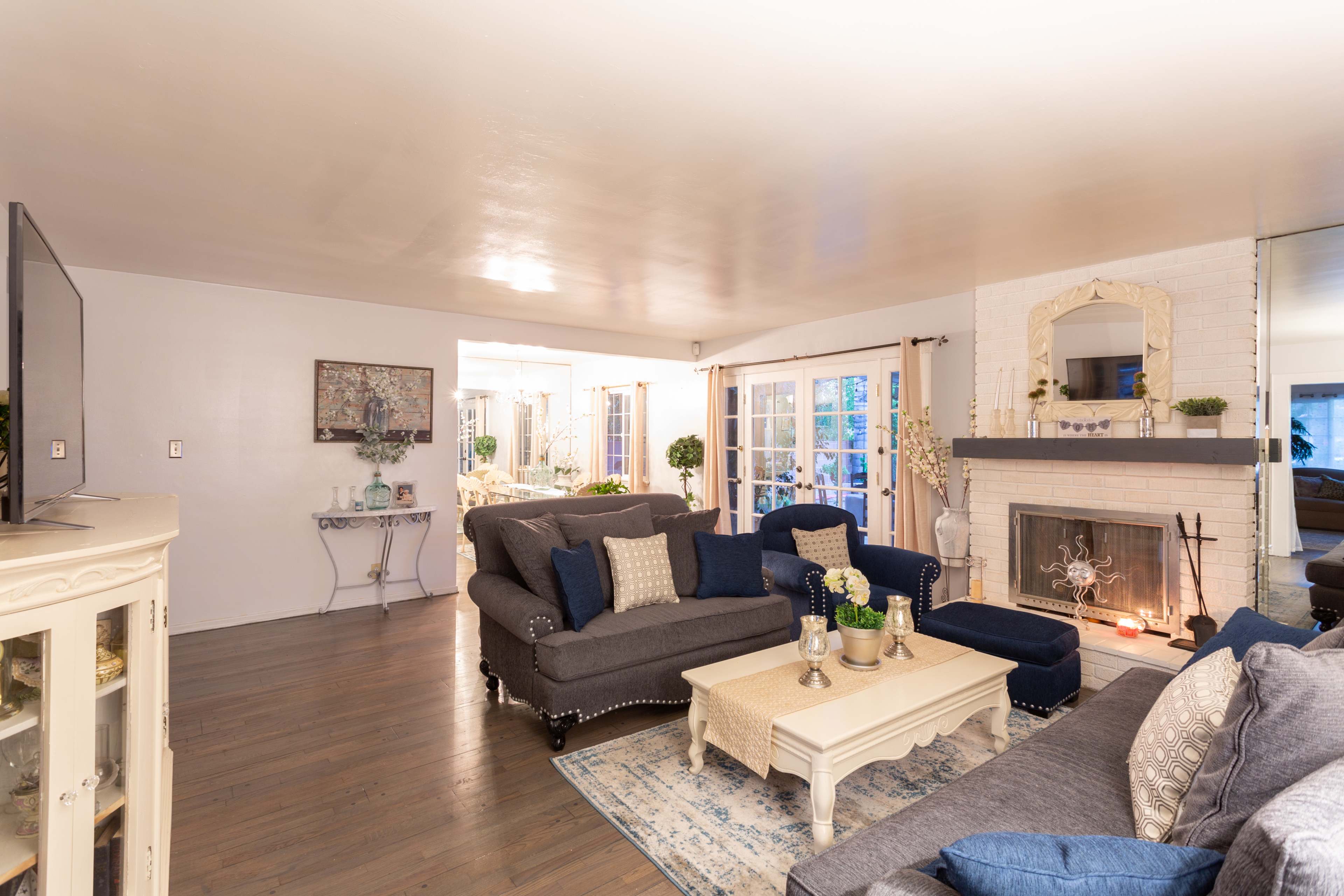 A cozy living room featuring a sectional sofa, a coffee table, and a fireplace with a mirror above it, leading to a bright space with French doors.