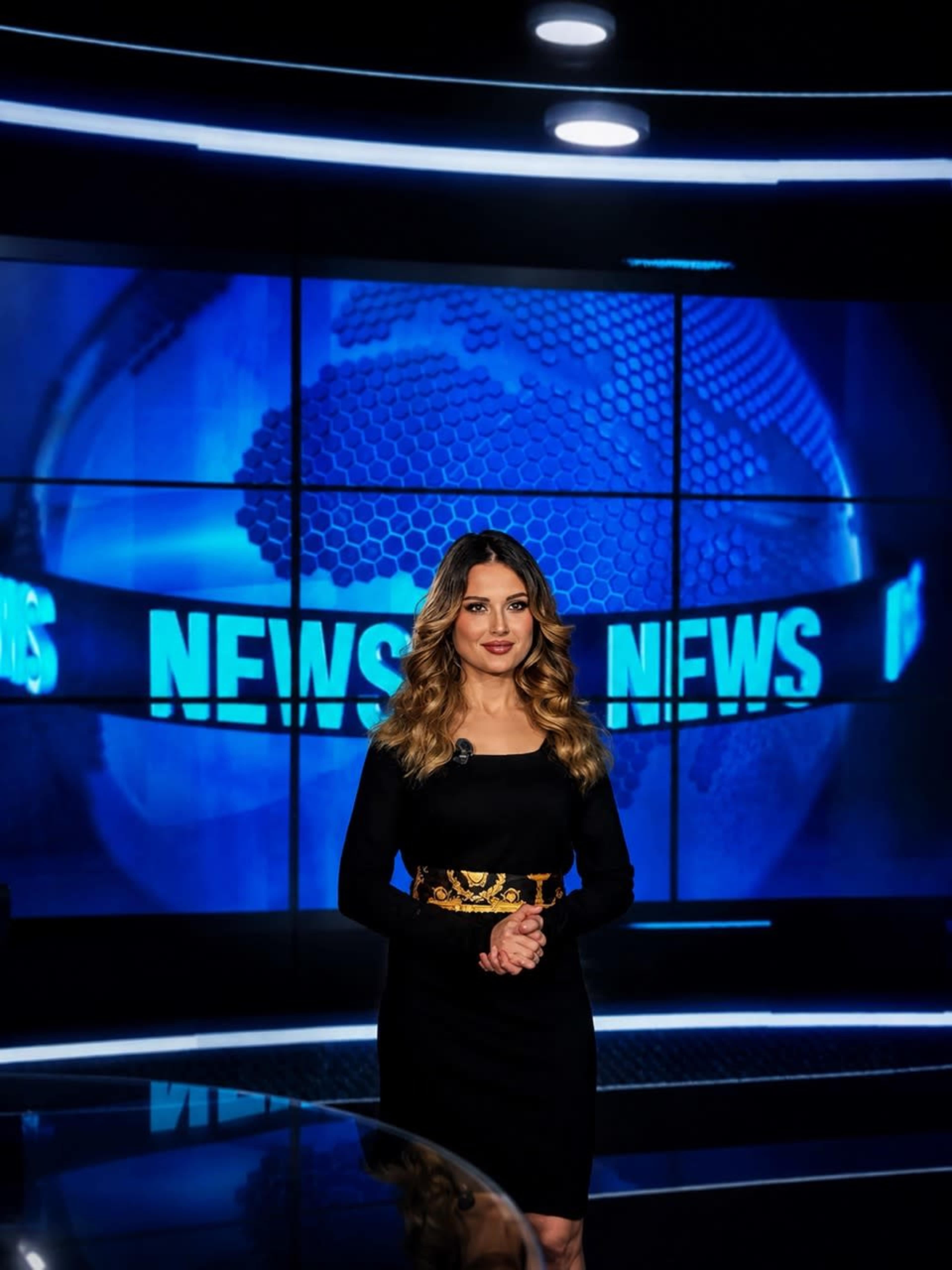 A news anchor stands in front of a large screen featuring the word "NEWS" in bold letters.