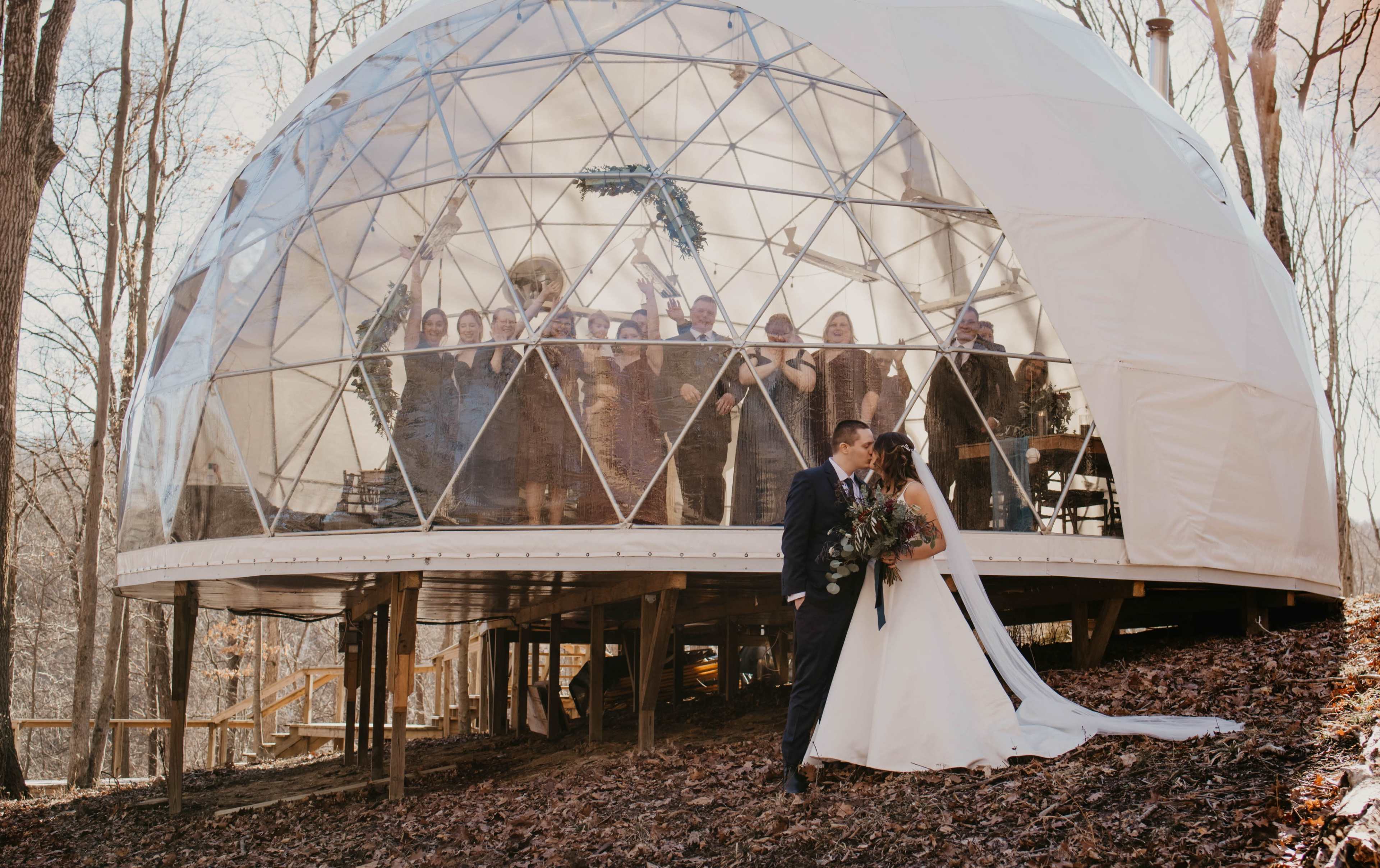 A couple shares a kiss in front of a geodesic dome venue, while guests celebrate inside.