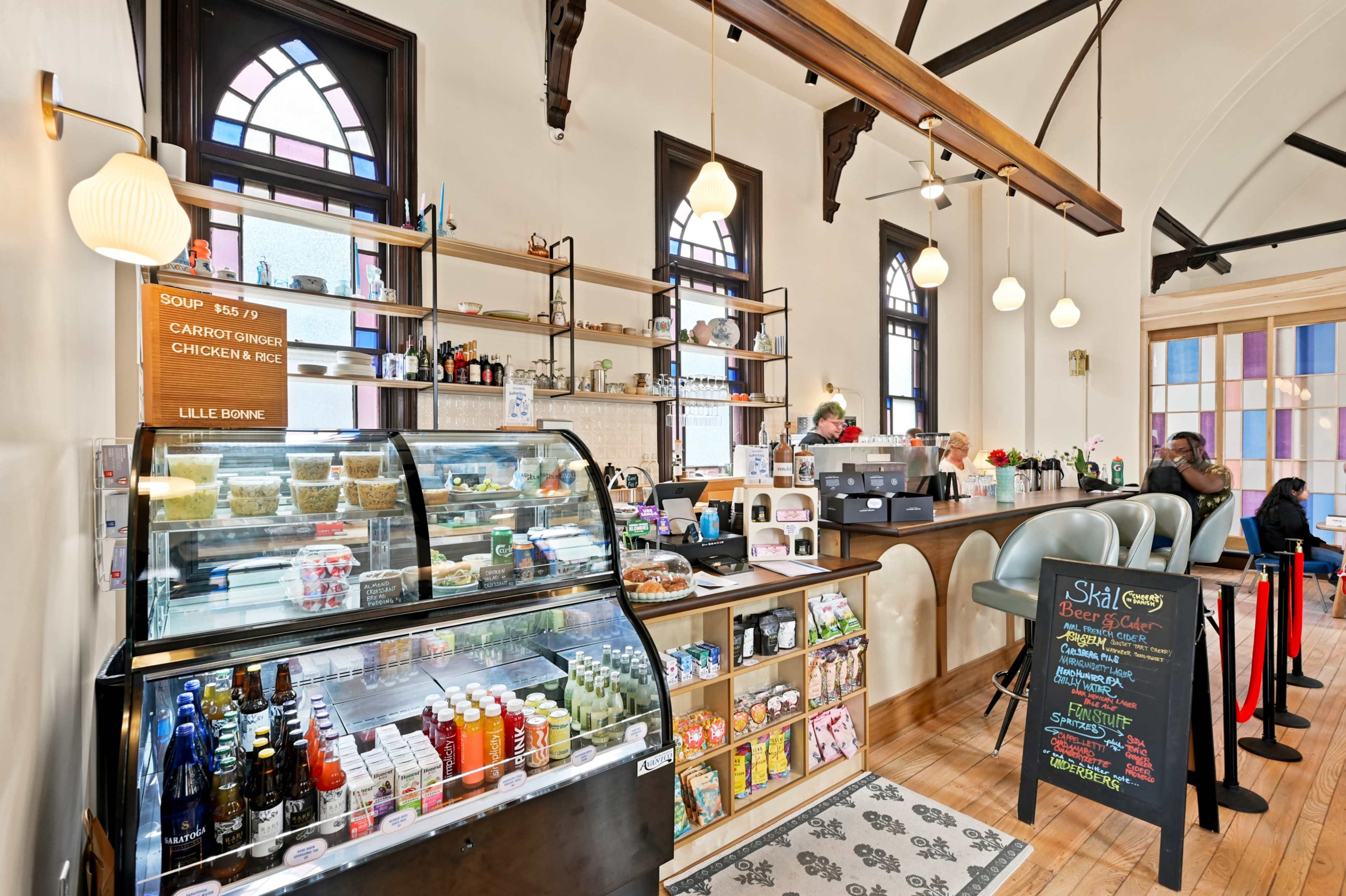 The image shows a cozy cafe interior with a display case of food and drinks, a service counter, and customers seated around the space.