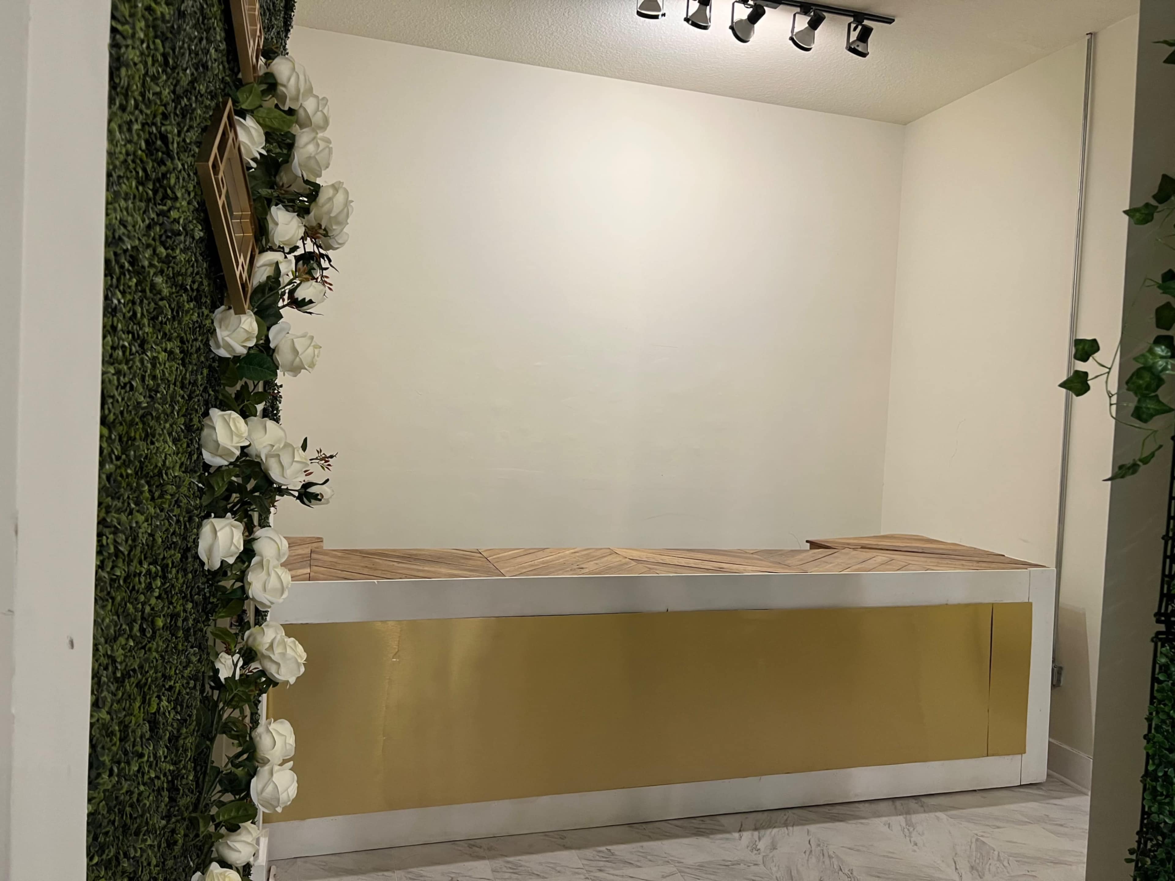 A reception area featuring a wooden counter with a gold accent, surrounded by artificial green foliage and white flowers.