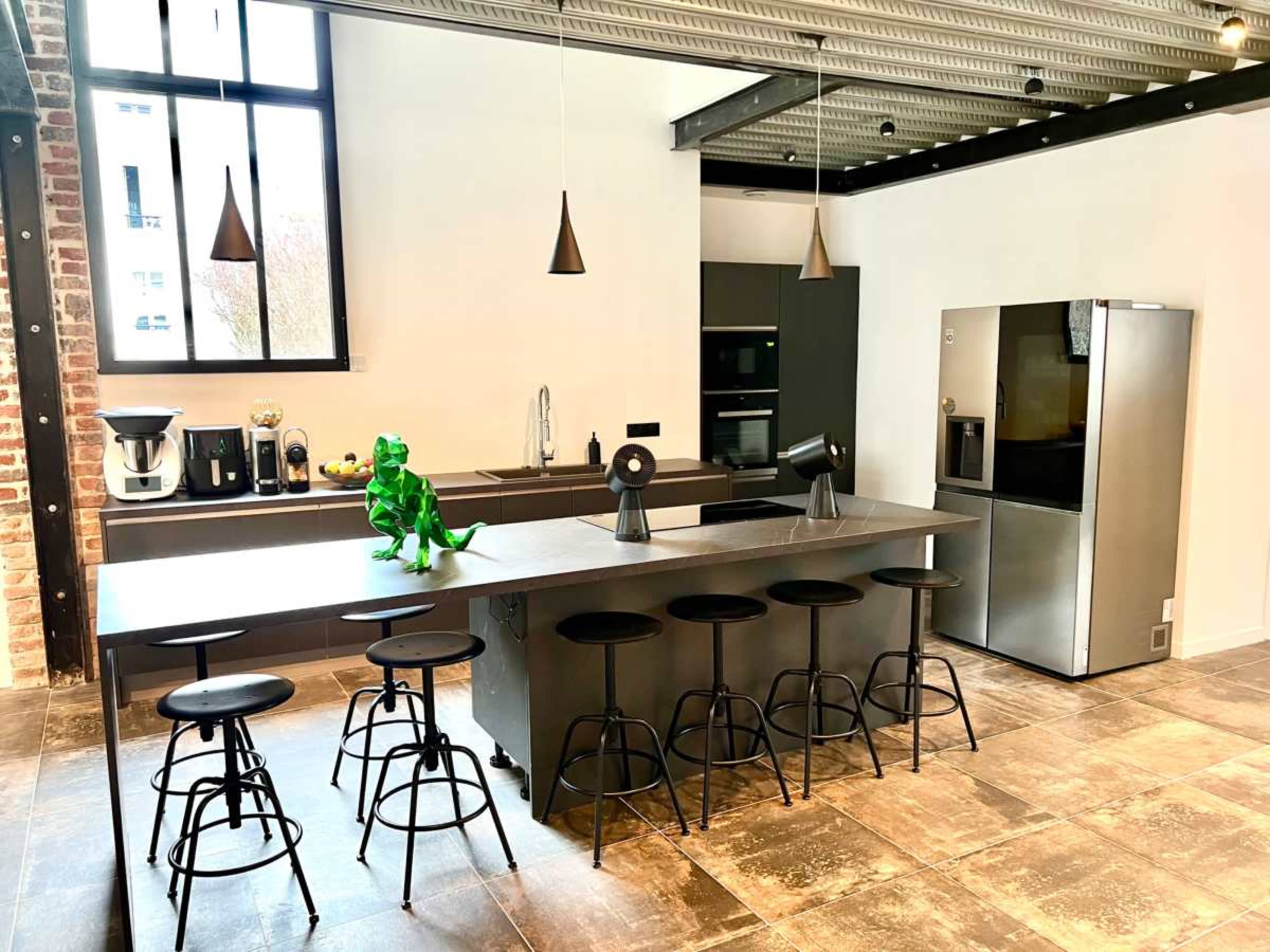 A modern kitchen features a large island with eight black stools, a stainless steel refrigerator, and a green decorative figure placed on the countertop.