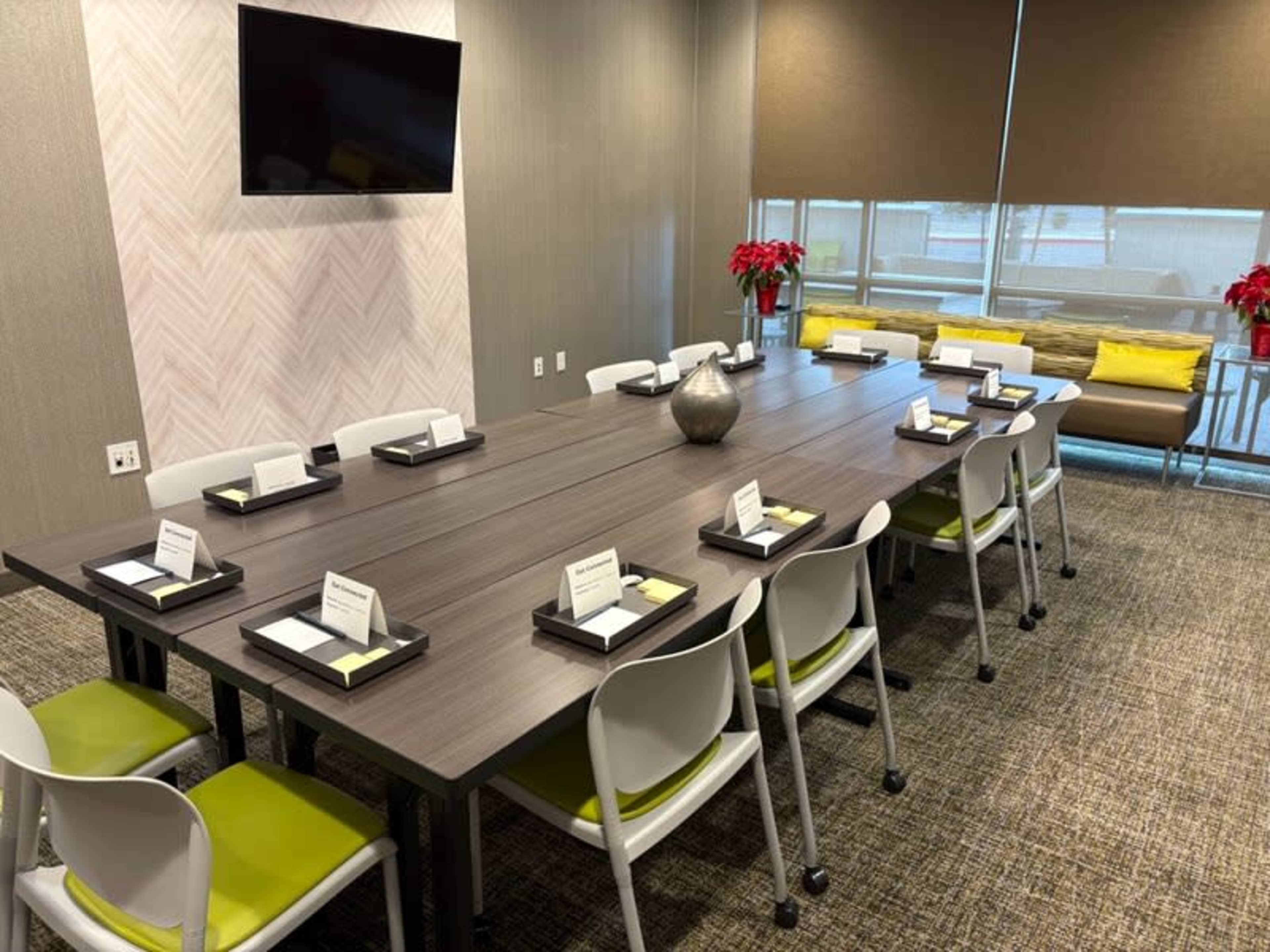 A conference room featuring a long table set with note pads and pens, surrounded by modern chairs, with a television mounted on the wall and natural light coming through large windows.