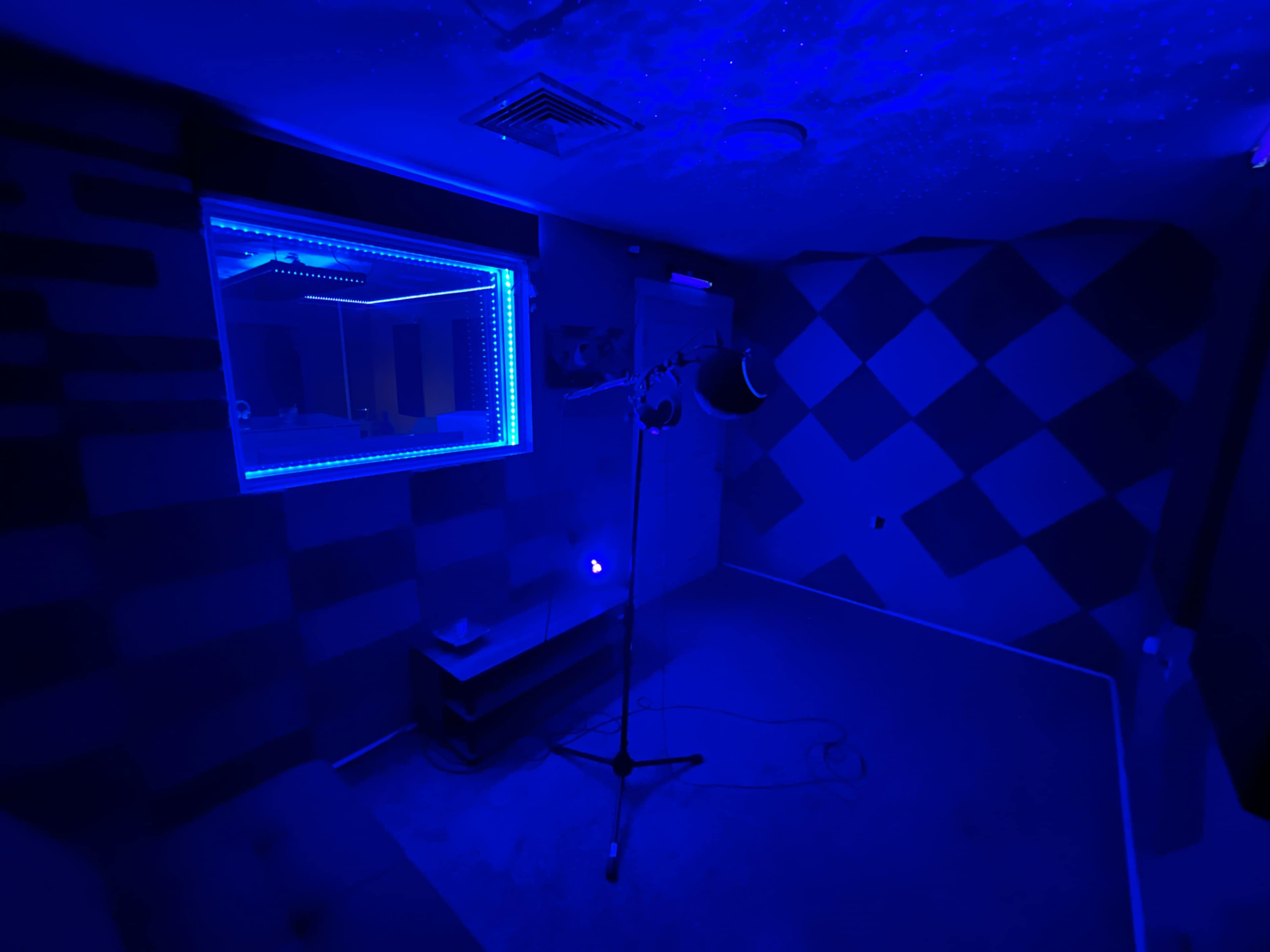 A small recording studio with blue lighting, a microphone on a stand, and a window with a view into an adjacent room.