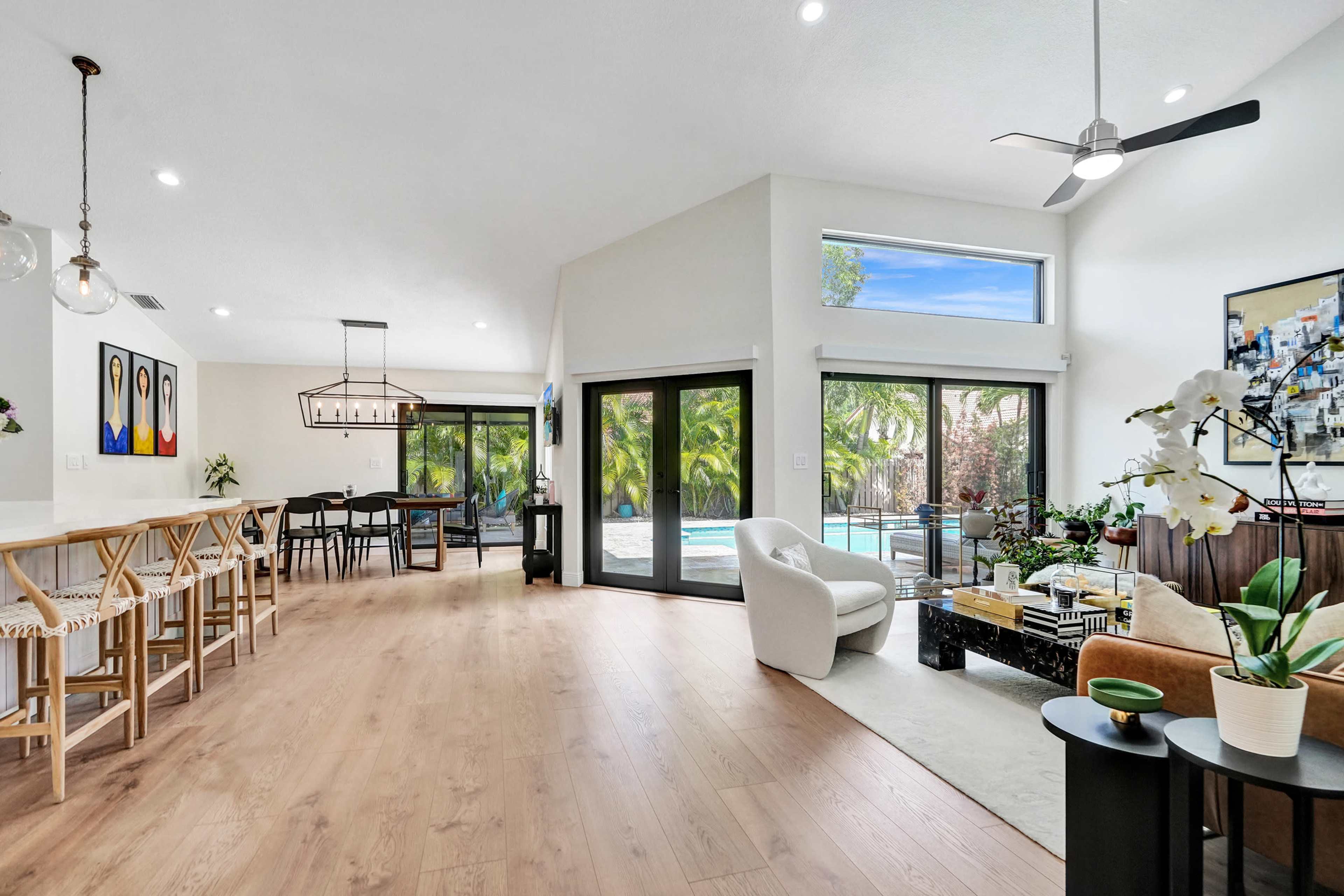 The image shows a bright, modern living space with a view of a pool and palm trees through large glass doors, featuring contemporary furniture and a dining area.