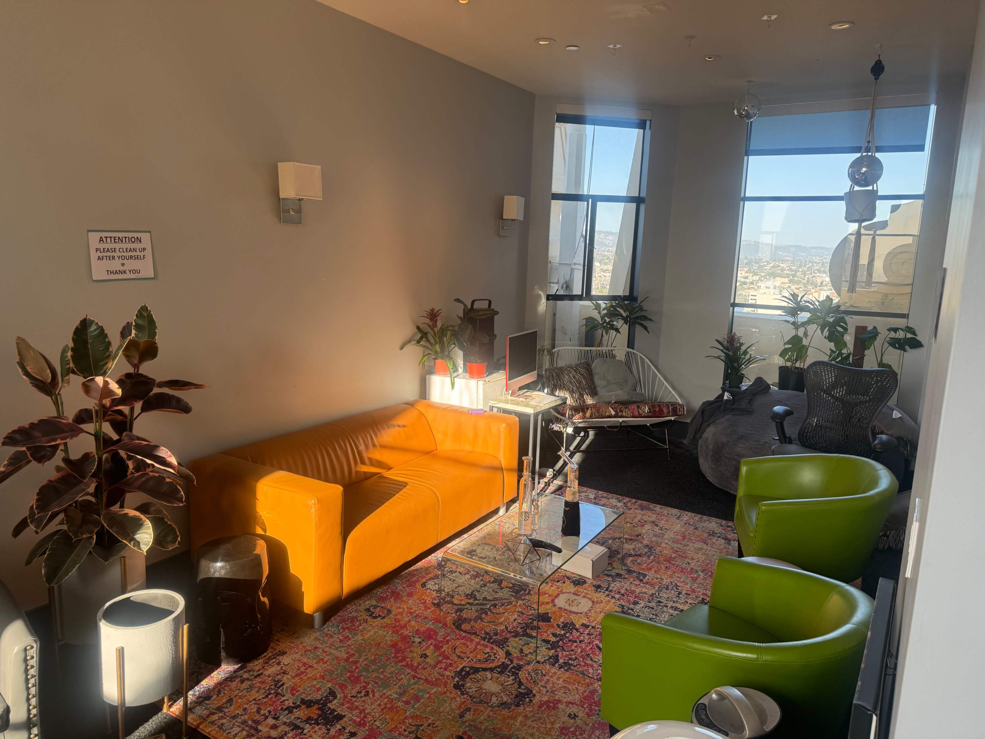 A bright room features an orange sofa, two green chairs, and a table with plants, along with a view of the city through large windows.