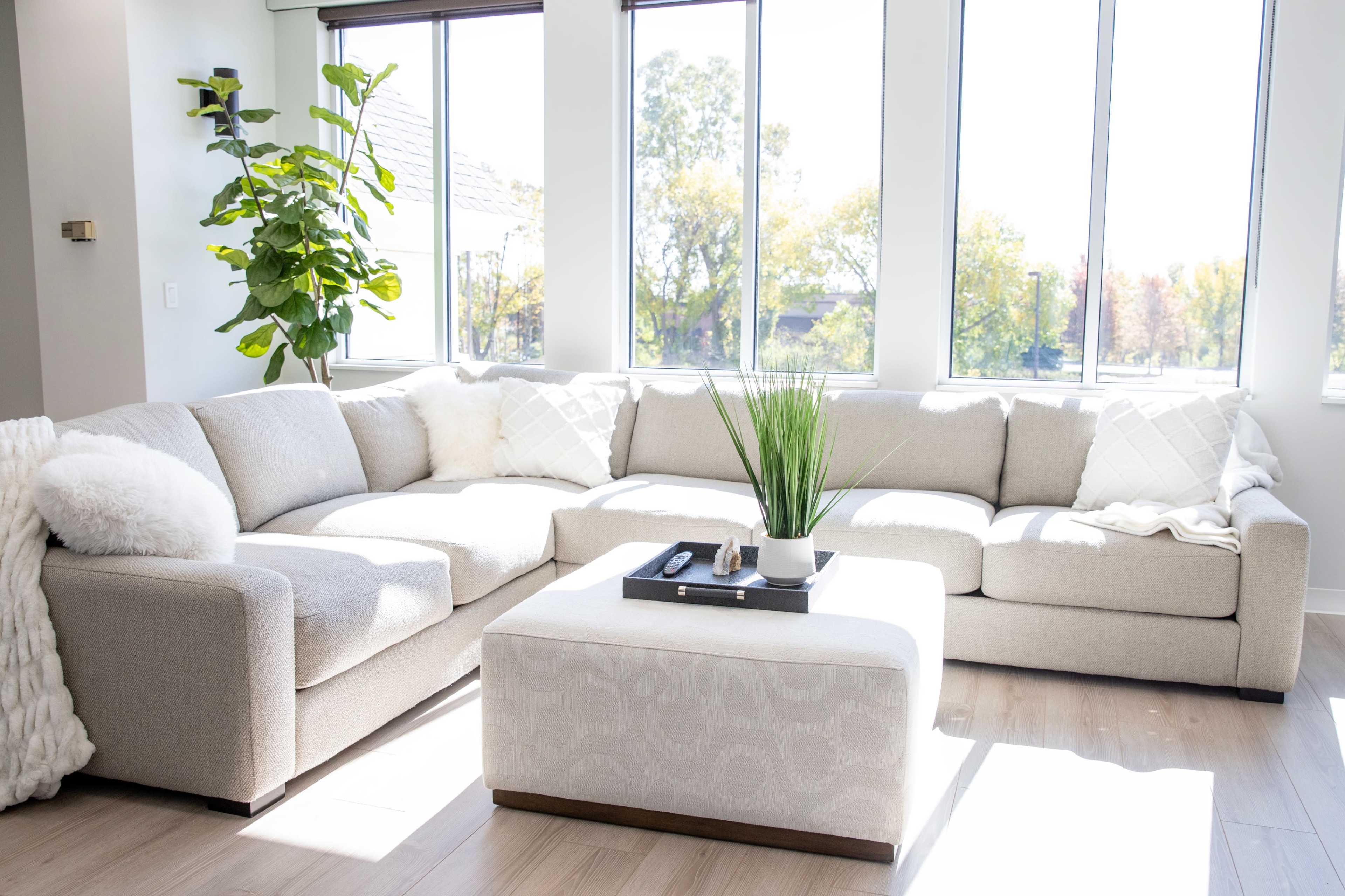 A spacious living room features a light gray sectional sofa and a matching ottoman, alongside large windows that allow natural light to illuminate the space.